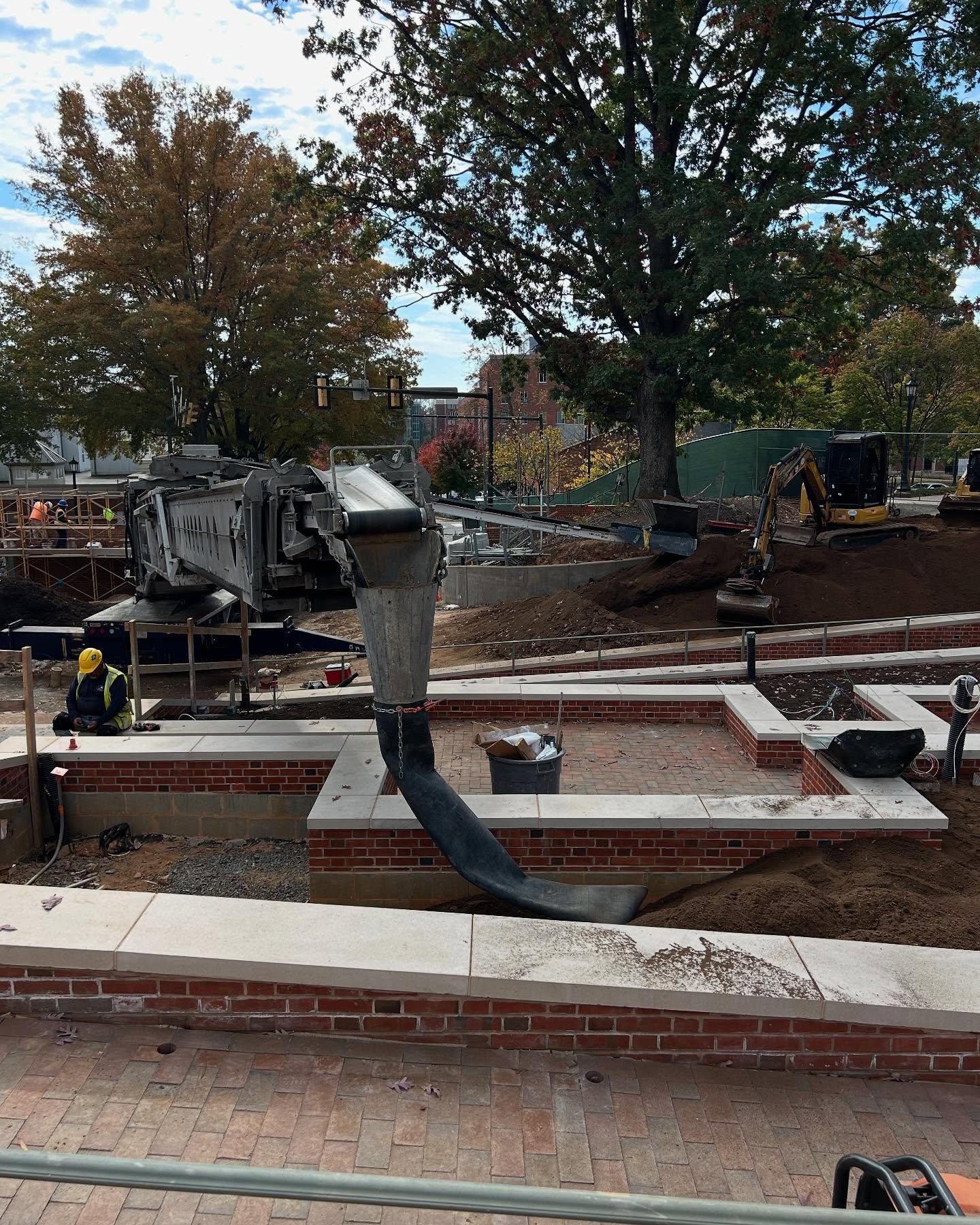 Construction site with workers, heavy machinery, and a large tree in the background. An industrial chute is pouring dirt into a dumpster.
