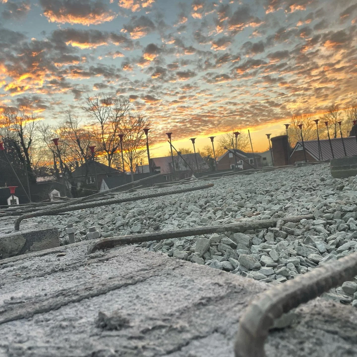 Low-angle view of a rooftop with gravel and metal bars, with a sunset sky filled with orange and gray clouds in the background, along with leafless trees and houses.