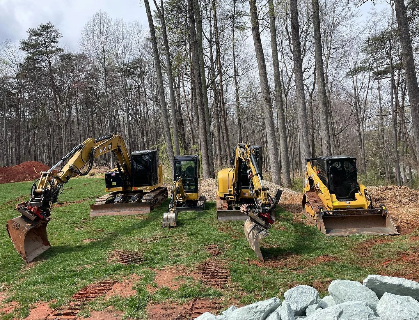 Four small Caterpillar excavators lined up on a grassy area in front of a wooded area during the day.