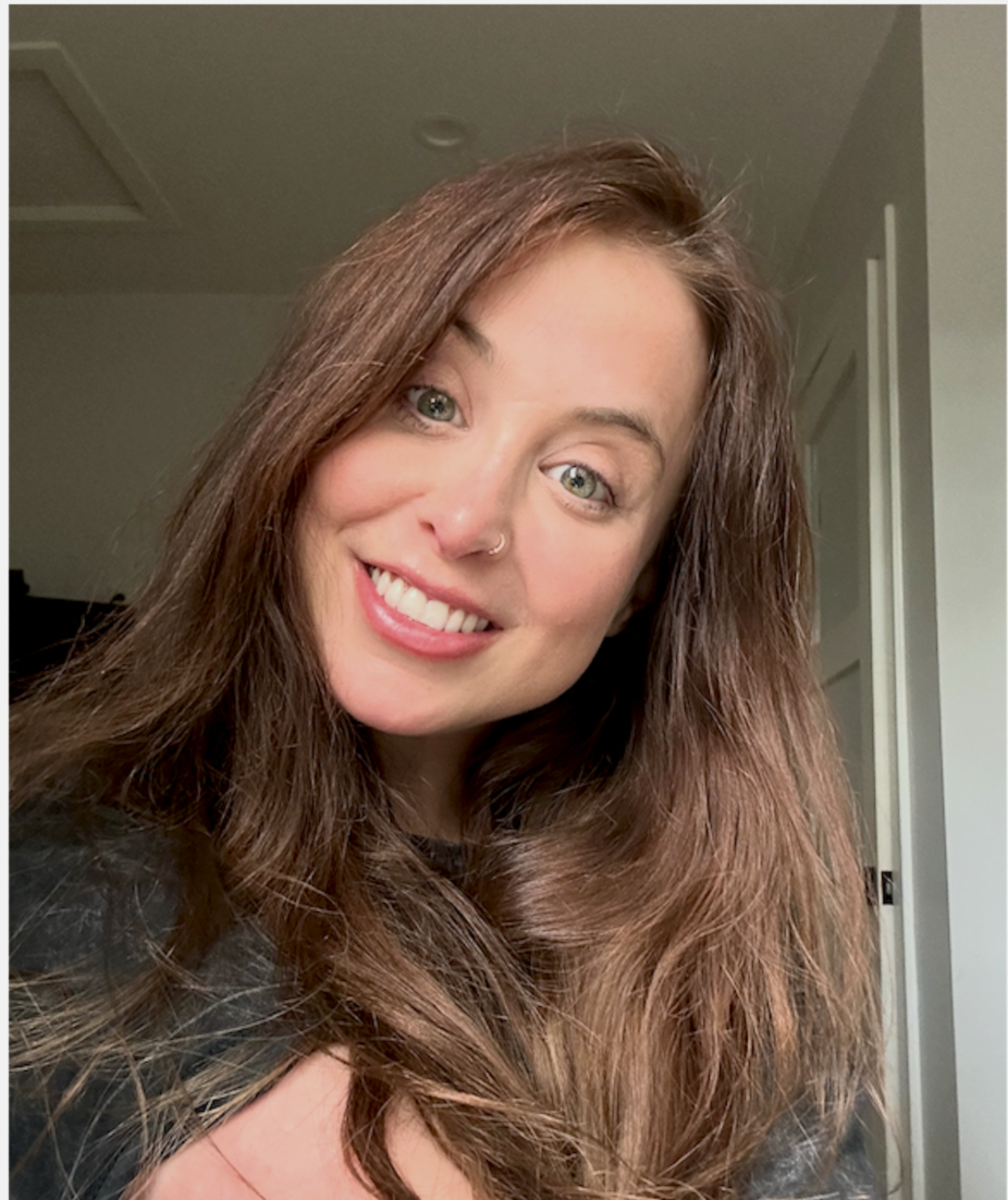 A woman with long, wavy brown hair, light skin, and a nose piercing smiling at the camera inside a home.