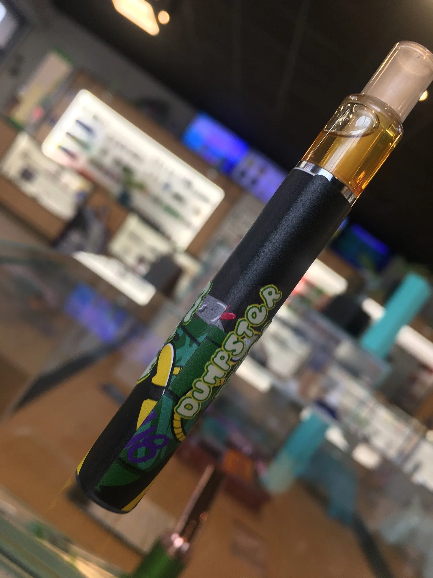 A vape pen with a colorful comic book style design featuring a green character and the words 'POW!'. The background shows a store interior with various items and screens.