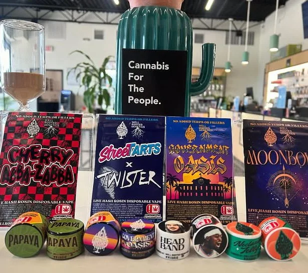 Display of cannabis-related products including a green cactus-shaped container with a sign that reads "Cannabis For The People," packets of flavored cannabis honey rosin in colorful packaging, and small containers with flavors like papaya, grape, and