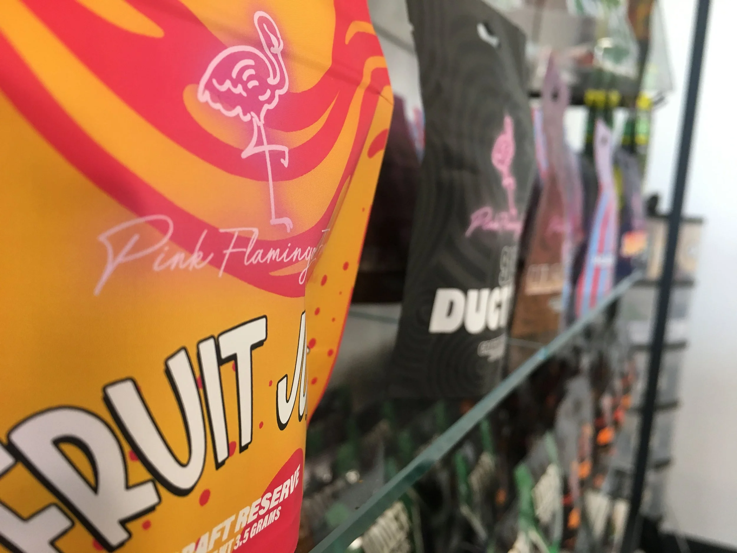 Close-up of a yellow bag of Flaming Hot Cheetos with a pink flamingo logo and the words "Pink Flamingo" on it, displayed on a glass shelf among other snack bags.