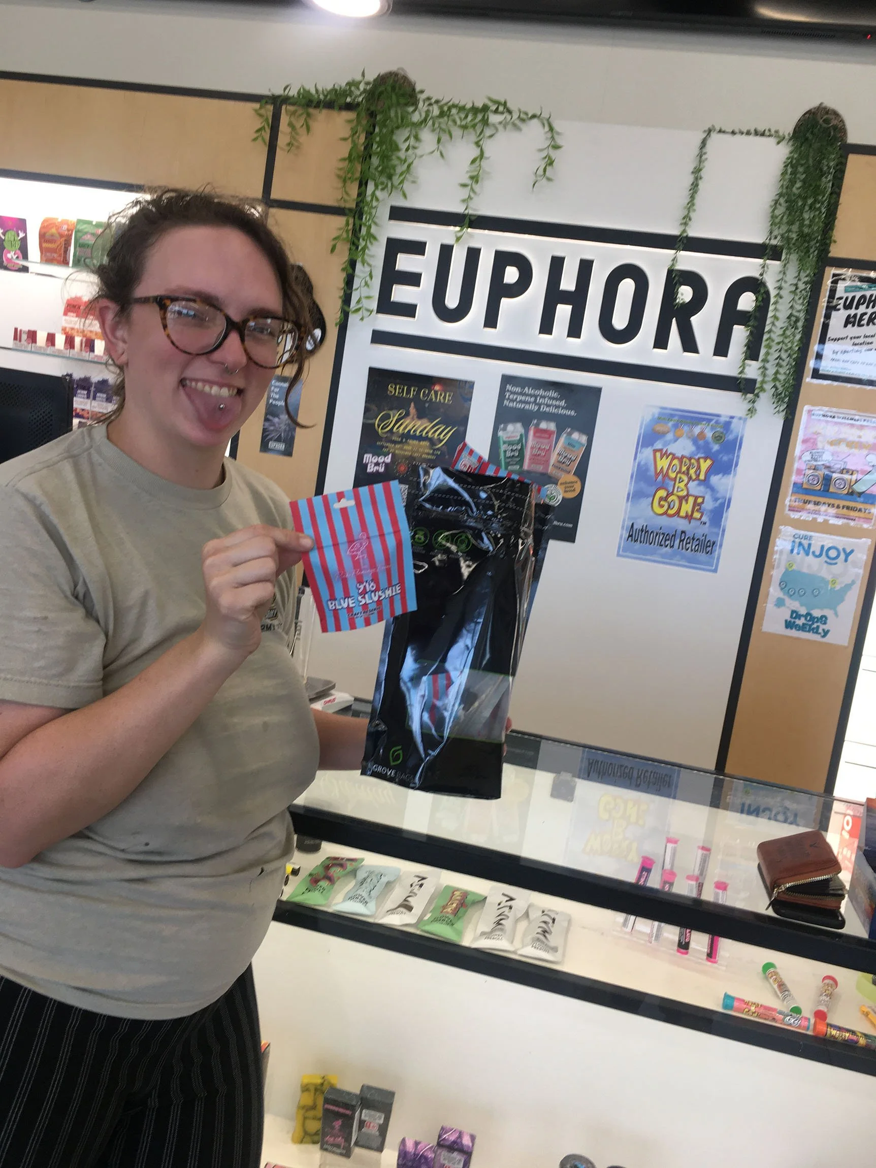 A woman with glasses and a lip piercing holding a striped red and blue candy bag and a package inside a store named EUPHORA, with candy and skincare products on display.
