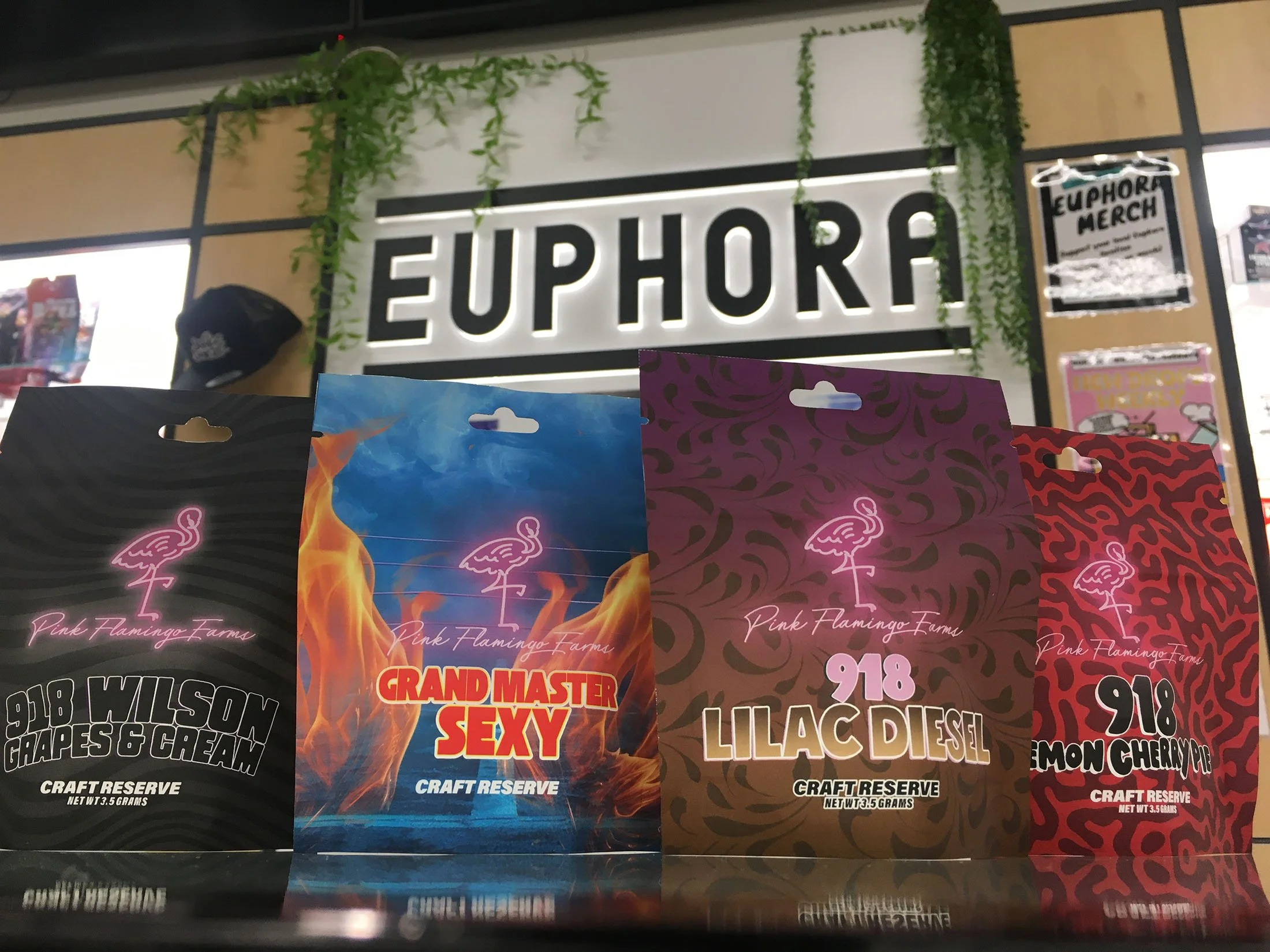 Colorful packaging of Pink Flamingo Farms craft reserve products displayed on a glass surface in front of a shop sign that reads 'EUPHORA', with green vines hanging above and a small poster on the wall to the right.