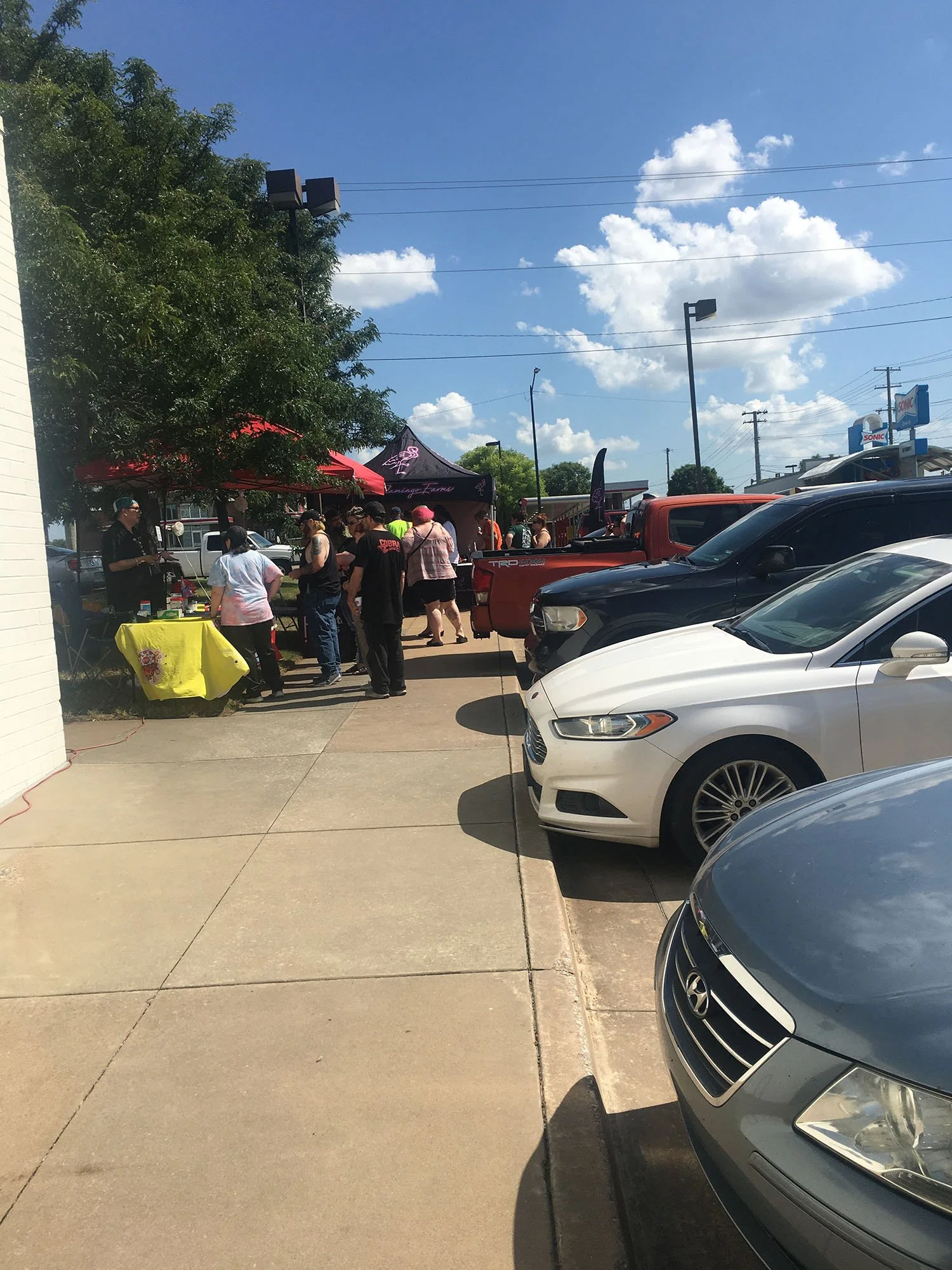 People gathered around outdoor market stalls, some under umbrellas, in a parking lot with cars parked along the curb, blue sky, and clouds.