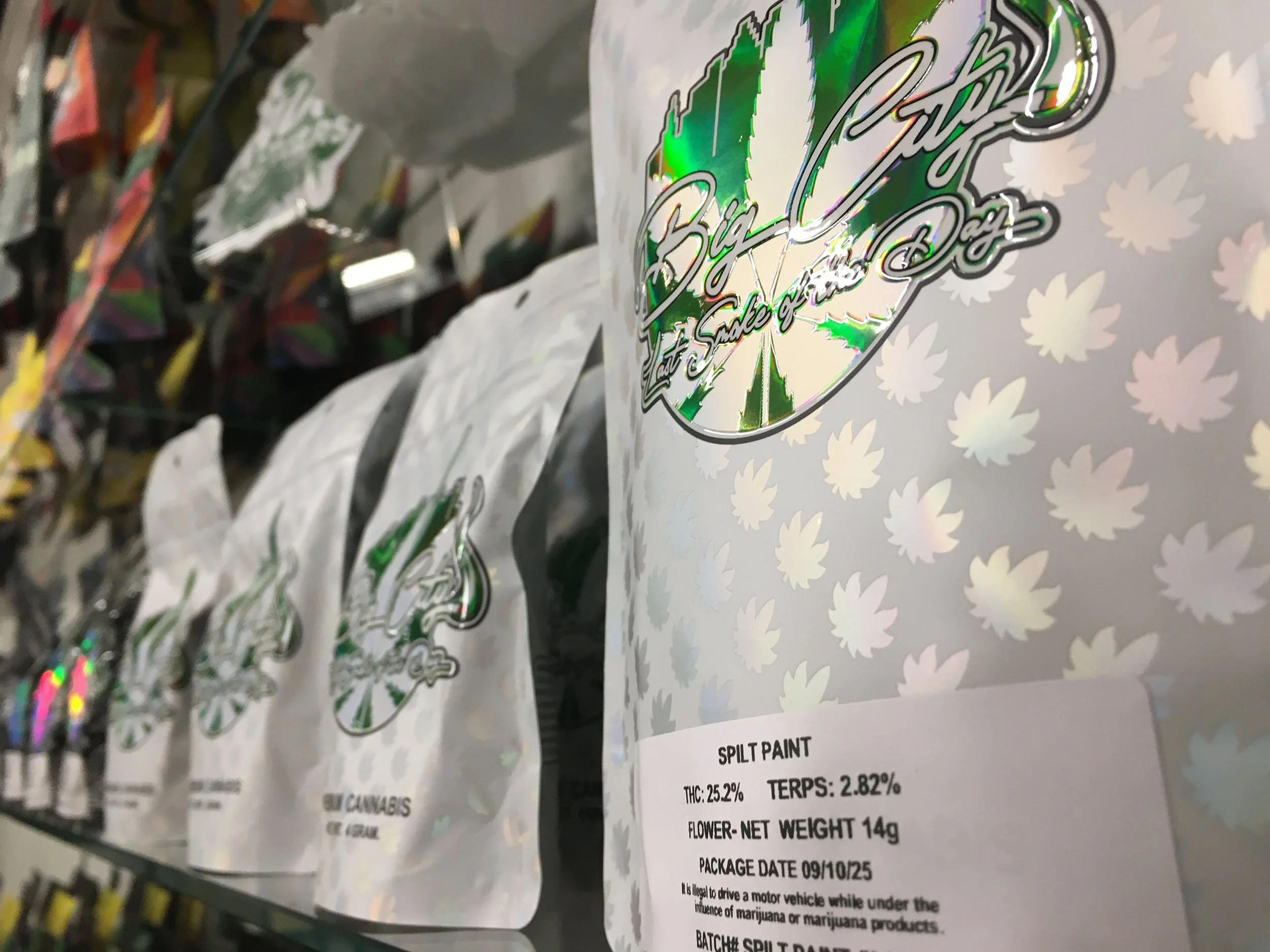 Holographic packaging for Big Cloud cannabis product, labeled 'Split Paint,' with THC 25.2%, terpenes 2.82%, and a flower net weight of 14 grams, on store shelf with other similar packages in the background.