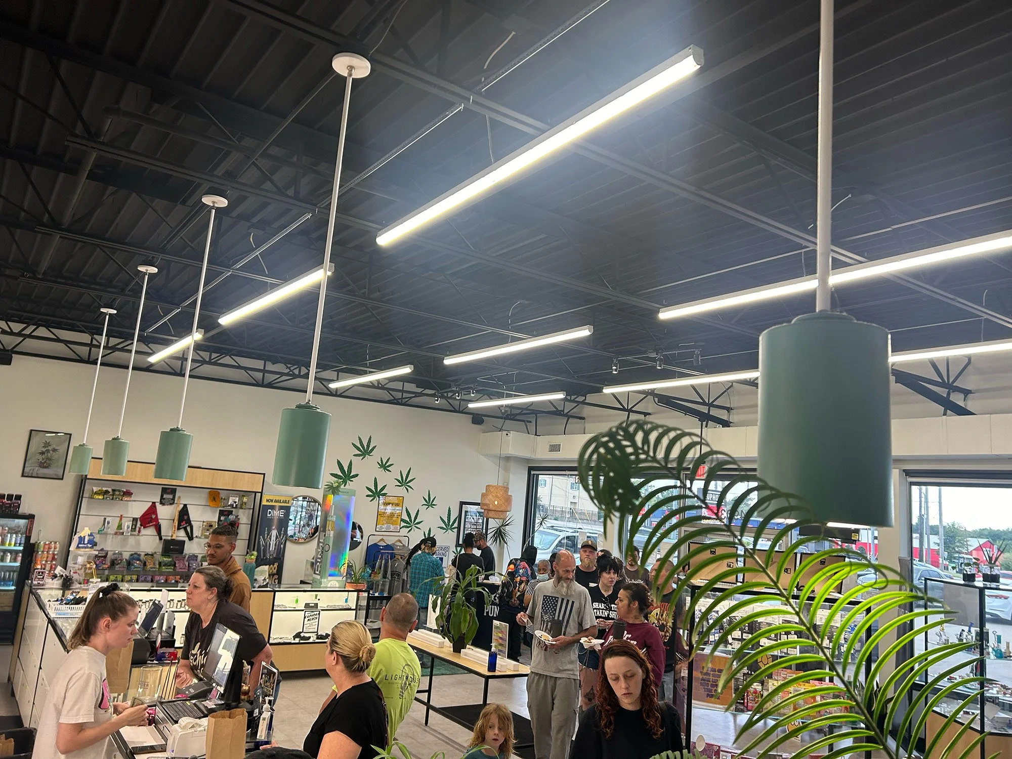 People standing in line and collecting orders at a counter inside a cannabis dispensary, with decorative marijuana leaf wall art, green hanging lights, and large windows.