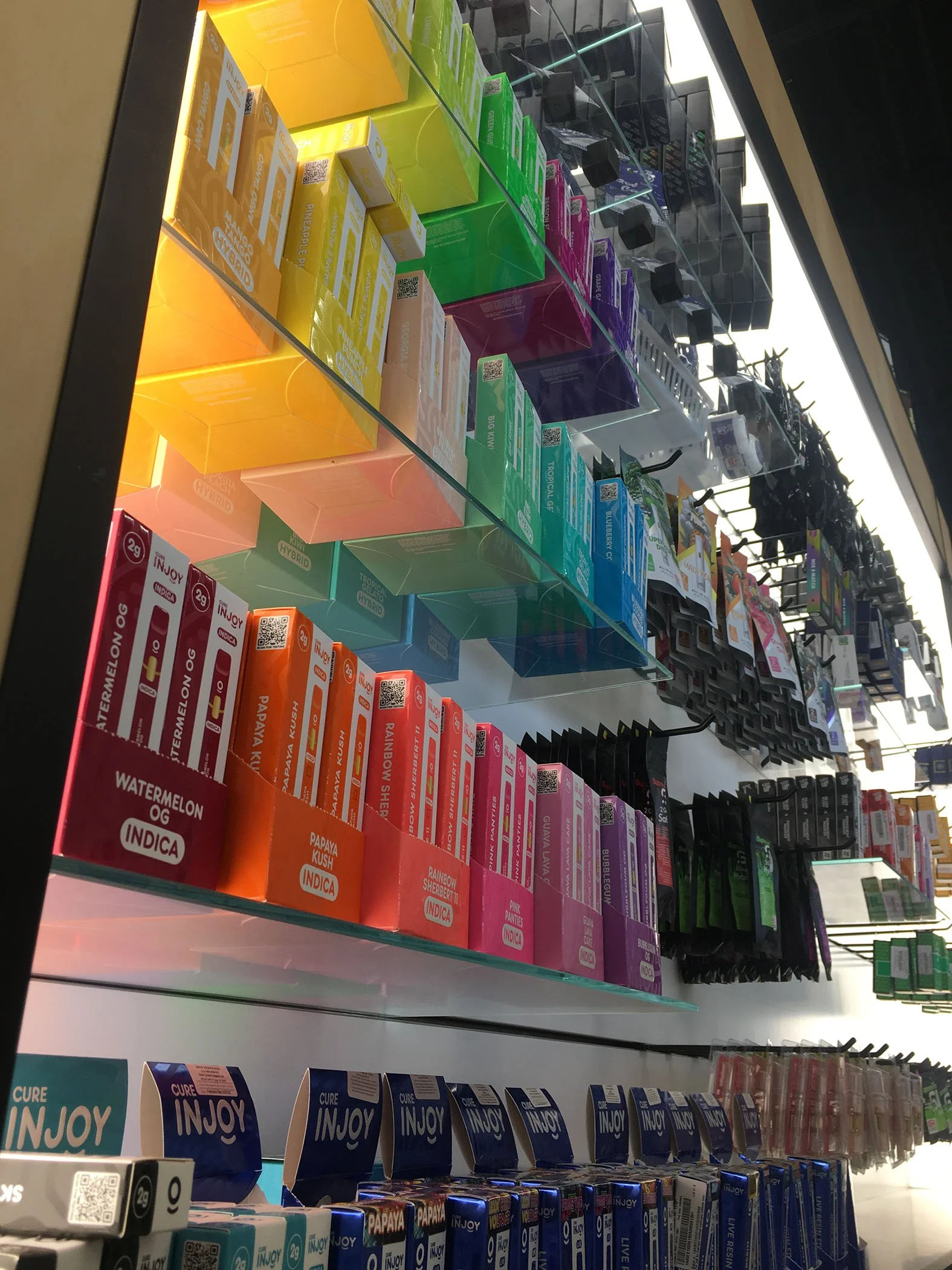 Display shelf of various colorful vape and smoking products, including boxes of e-liquids in different flavors and sizes, arranged in rows in a retail store.