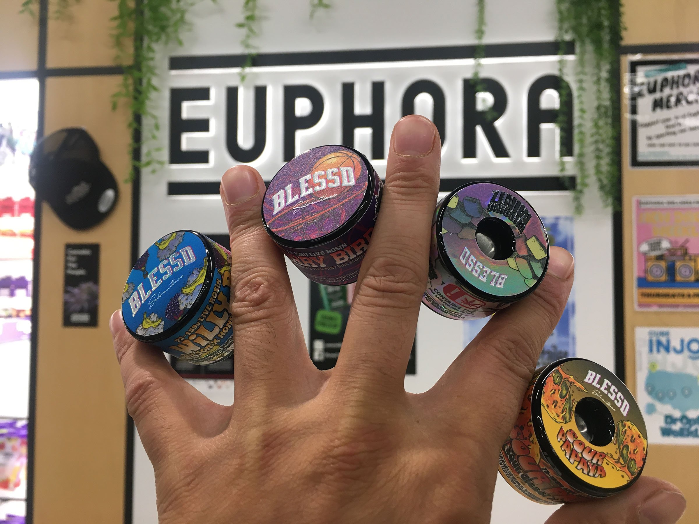 A person's hand holding various small containers of Blissed Superfood in front of a store sign that reads 'Euphoria' with hanging green plants above.