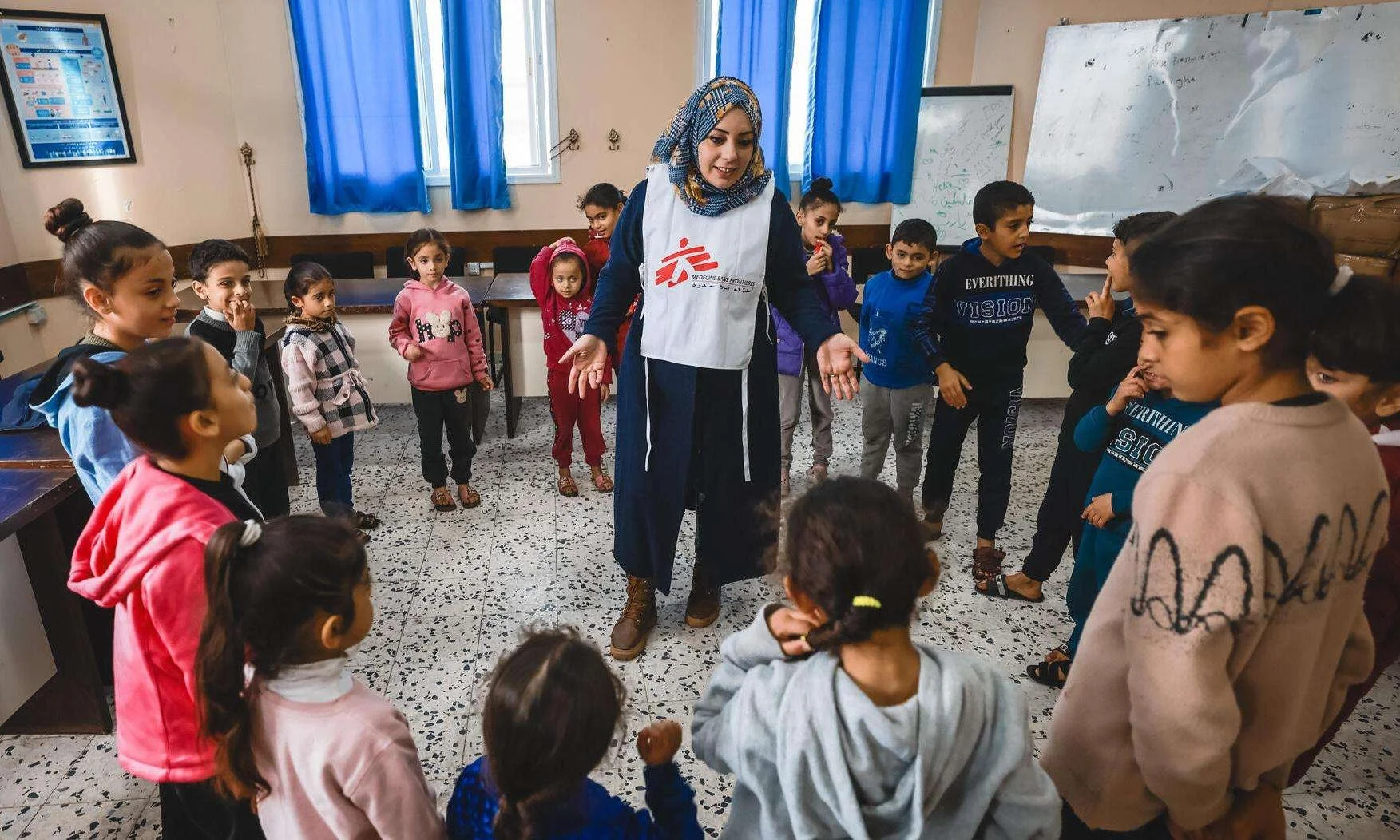 A woman wearing a vest with a red logo stands in the middle of a group of children in a classroom, engaging in a group activity.