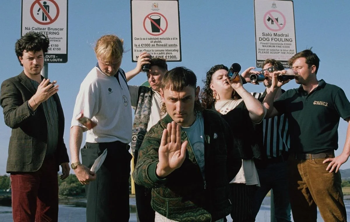 Group of young people standing outdoors in front of blue sky, some looking at phones or cameras, one holding beer bottles, with signs in Irish about prohibitions, including no smoking, no drinking, and no dog fouling.