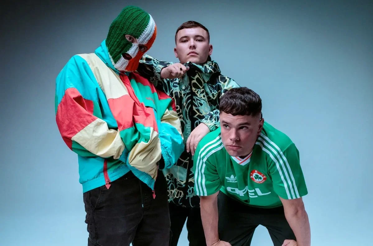 Three young men with serious expressions posing against a plain background. One wears a colorful windbreaker and a ski mask, another wears a patterned jacket, and the third wears a green Adidas sports jersey.