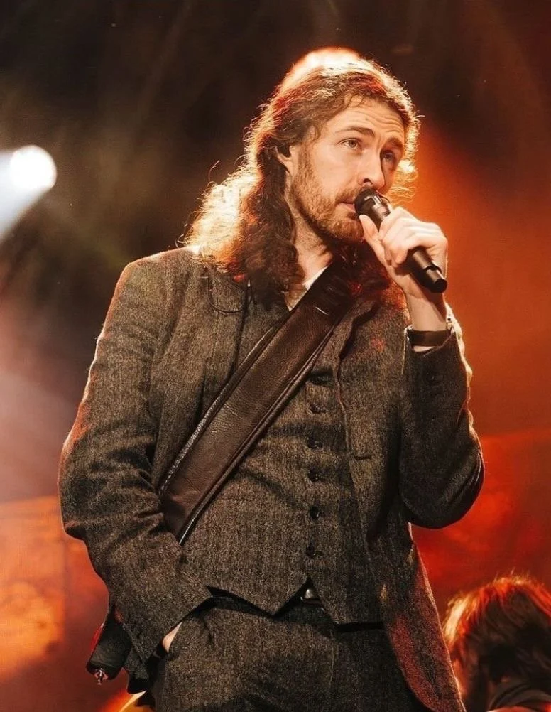 A man with long hair and a beard holding a microphone on stage, wearing a brown suit with a crossbody bag, performing under warm lighting.