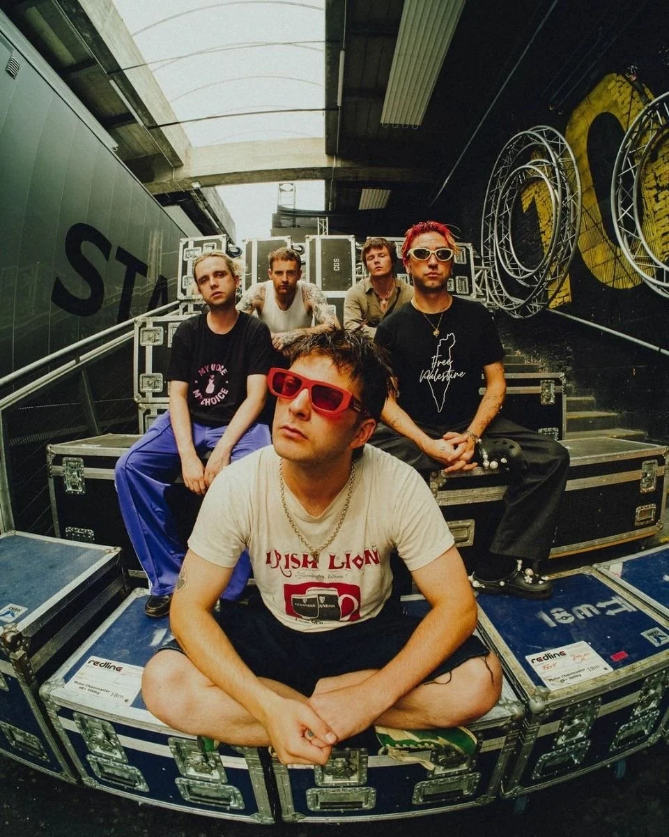 A group of five young men sitting on stage equipment cases in a backstage corridor, with musical equipment and circular metal stage elements behind them.