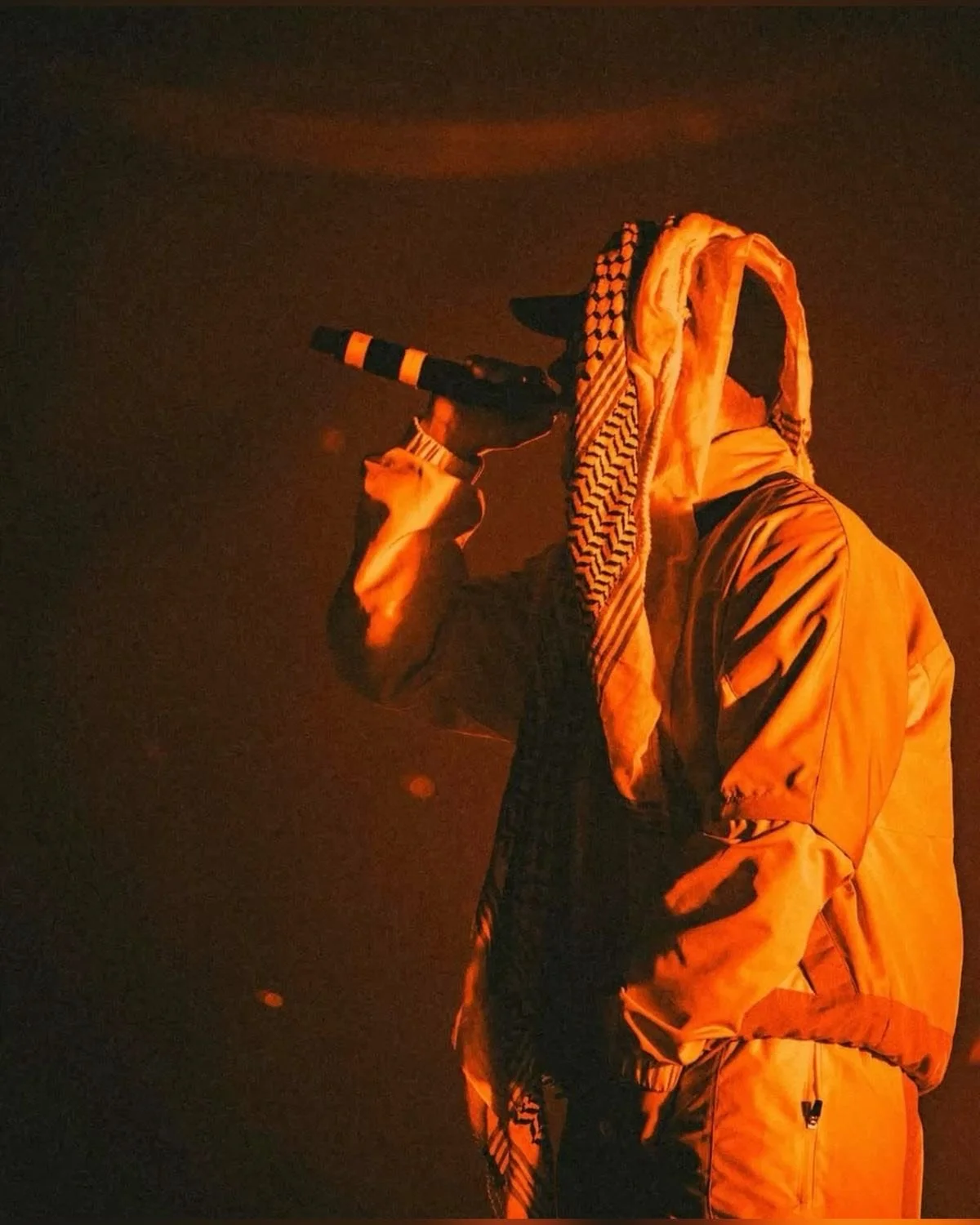 Person wearing light-colored clothing, including a jacket and headscarf, holding a camera or binoculars to their eyes, with a dark background and orange lighting.