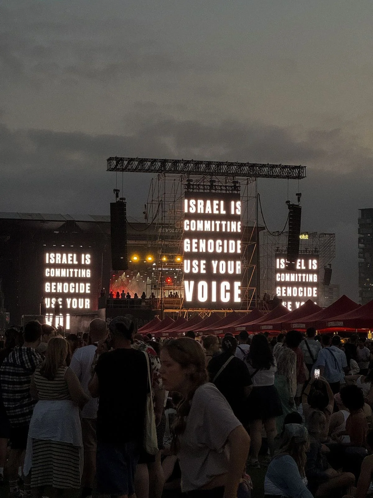 Large electronic screens display an anti-violence message at a gathering or protest, with a crowd of people in front of tents and a stage in the background.