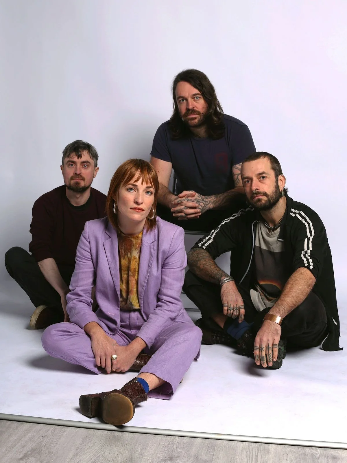 Group of five people, with one woman in a lavender suit sitting on the floor in the foreground, and four men, one kneeling behind her and three sitting on the floor, against a plain white backdrop