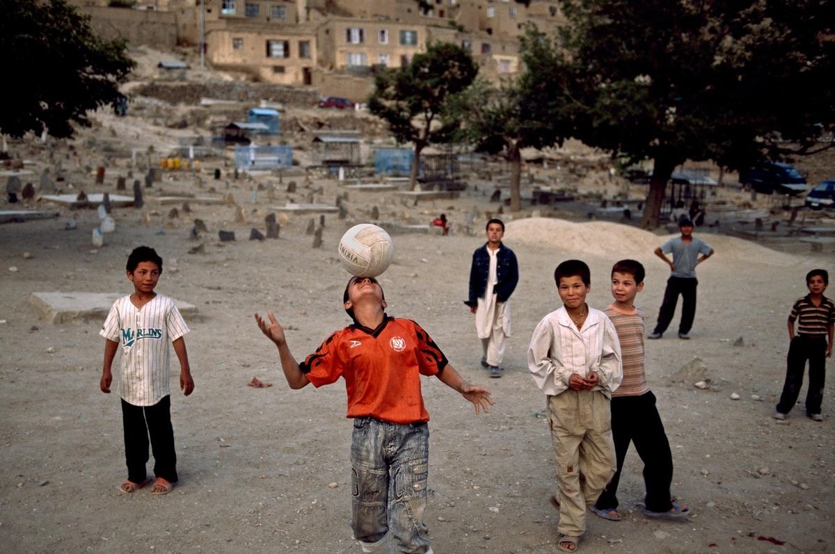 Children playing with a soccer ball in a dusty outdoor area with some buildings and trees in the background.