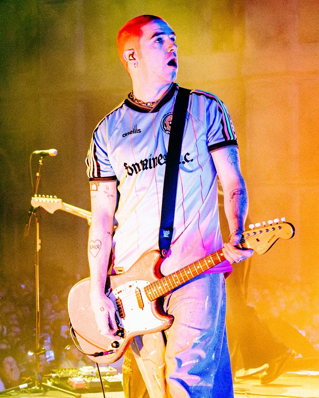 A young male musician with tattoos, wearing a soccer jersey, playing an electric guitar on stage with colorful lighting.