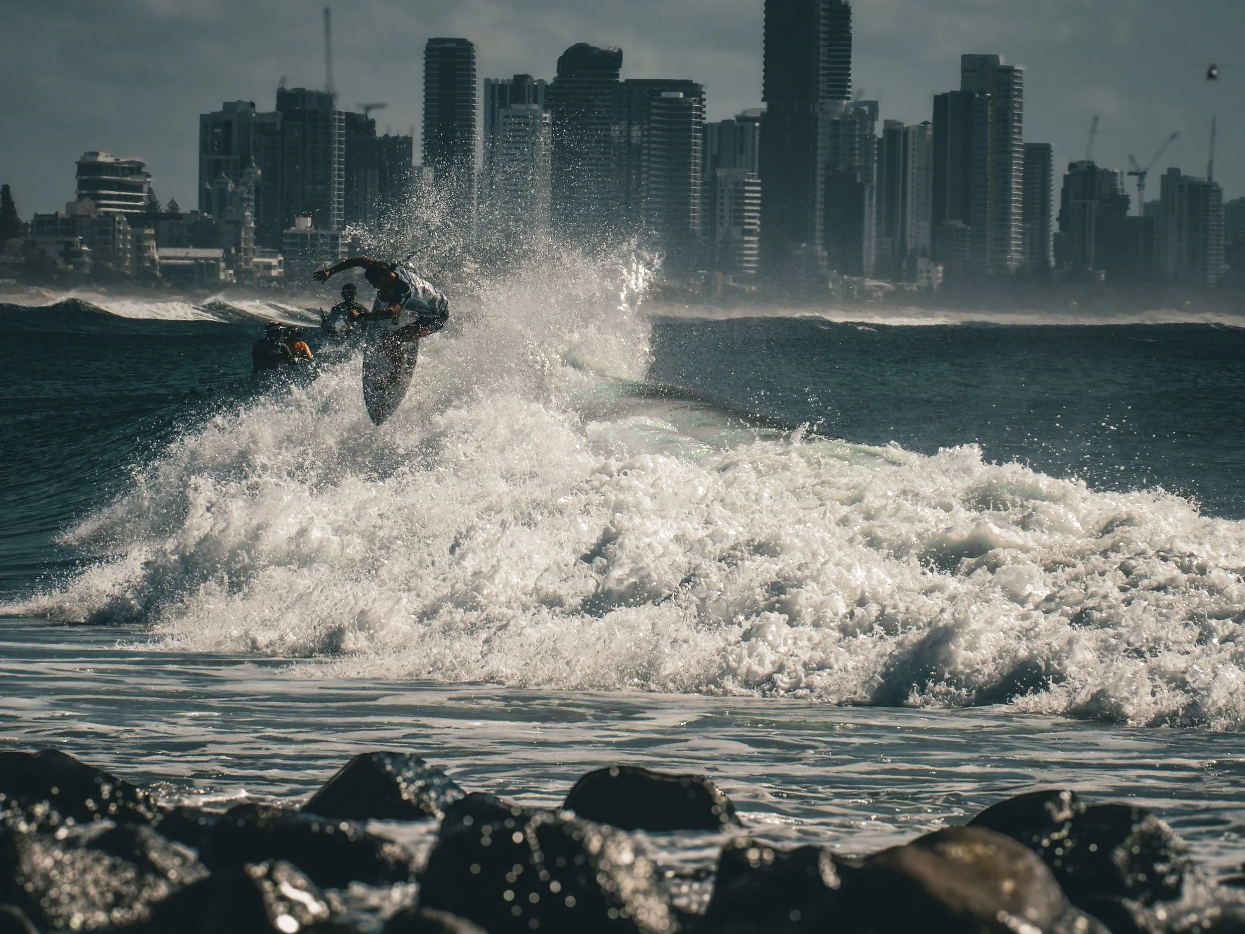 A person surfing on a wave in an urban beach setting with city skyscrapers in the background.