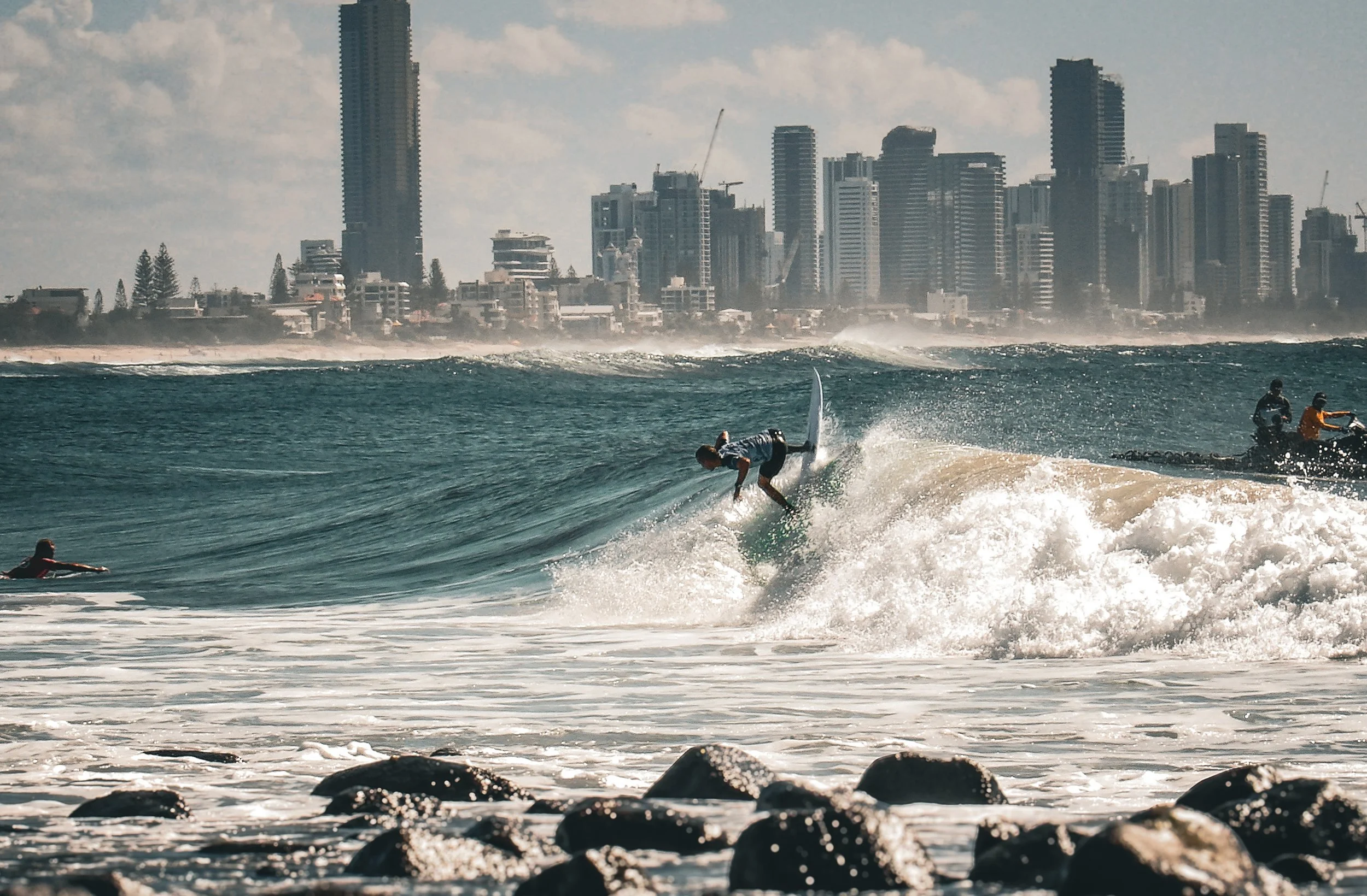 A person surfing on a large wave near rocks on the beach, with a city skyline of tall buildings in the background.