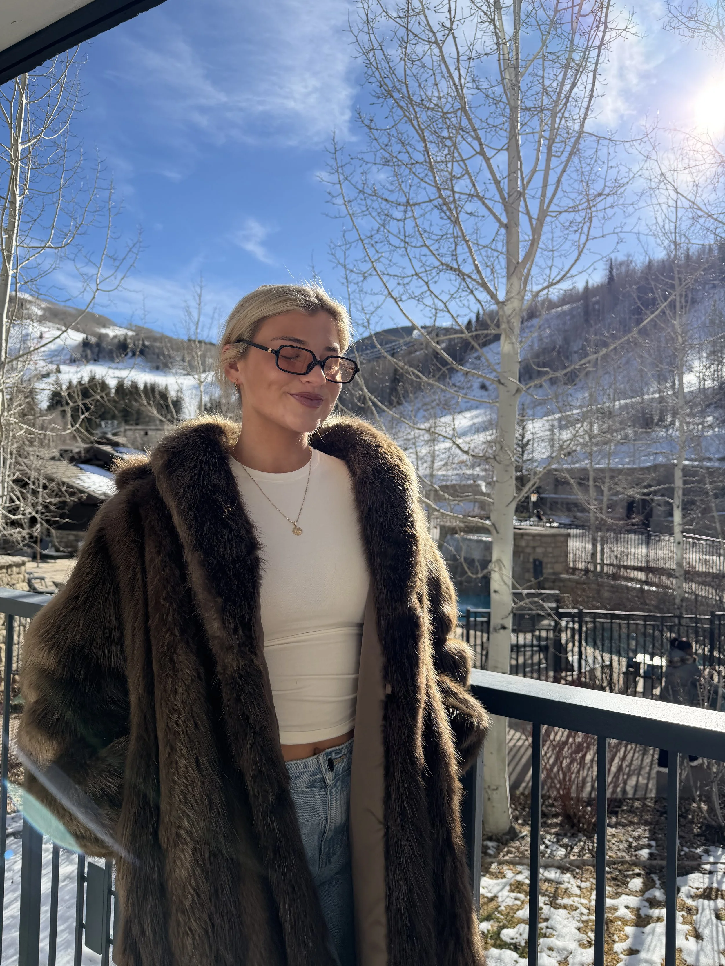 A woman with blonde hair wearing glasses, a white crop top, a fur coat, and jeans stands outdoors on a fence balcony during winter, with snow-covered hills and leafless trees in the background under a clear blue sky.
