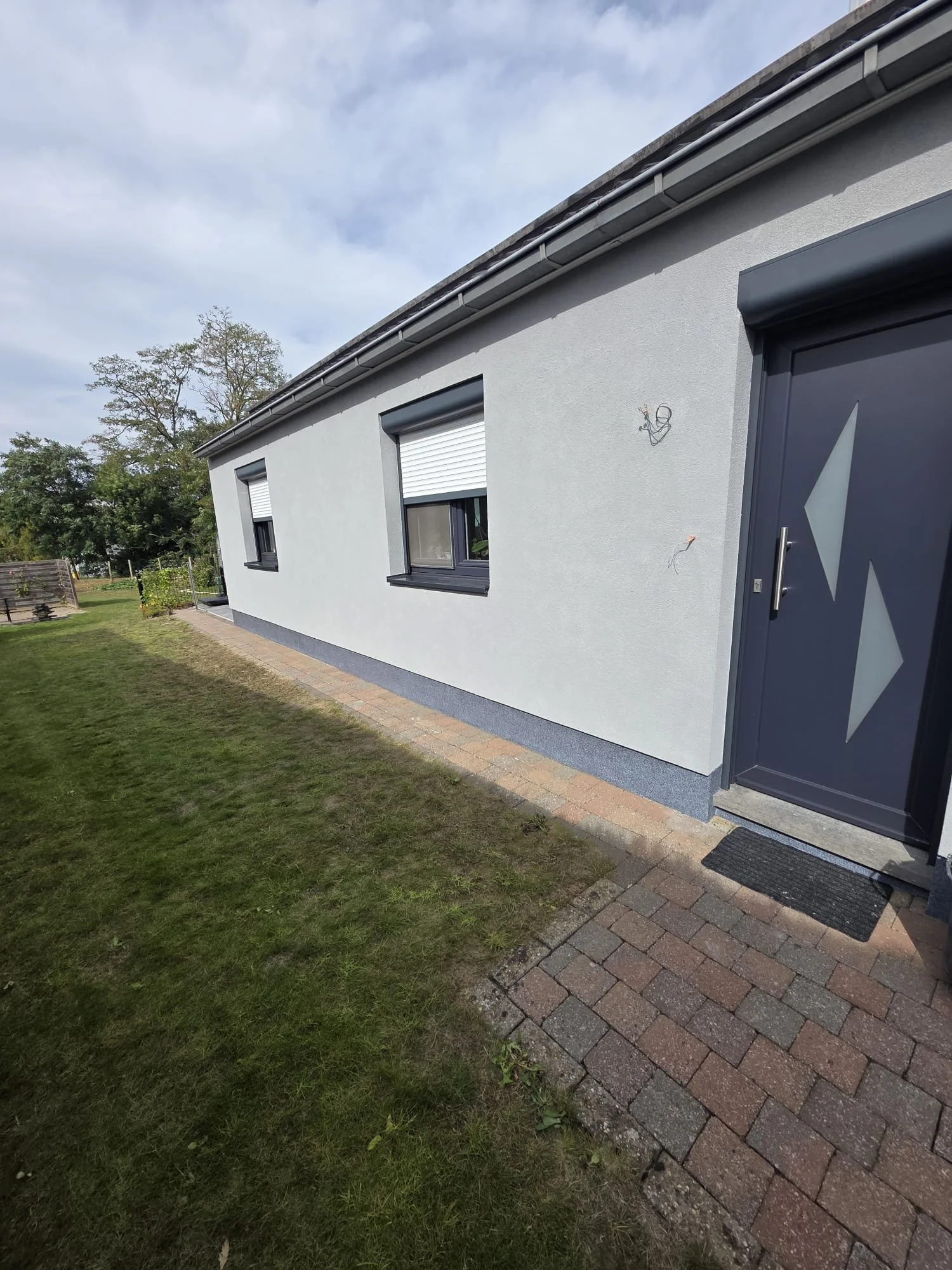 Front of a modern house with two black window frames, gray exterior wall, a gray door with decorative glass, and a brick walkway bordered by grass, under a cloudy sky.