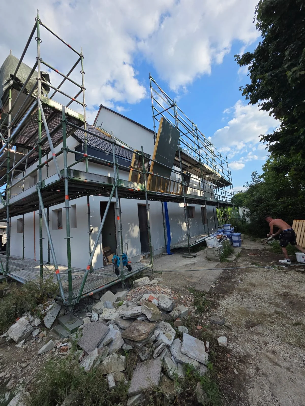 Construction site of a building with scaffolding surrounding the white exterior walls, some construction materials on the ground, a worker working on the ground, and a partly cloudy sky overhead.