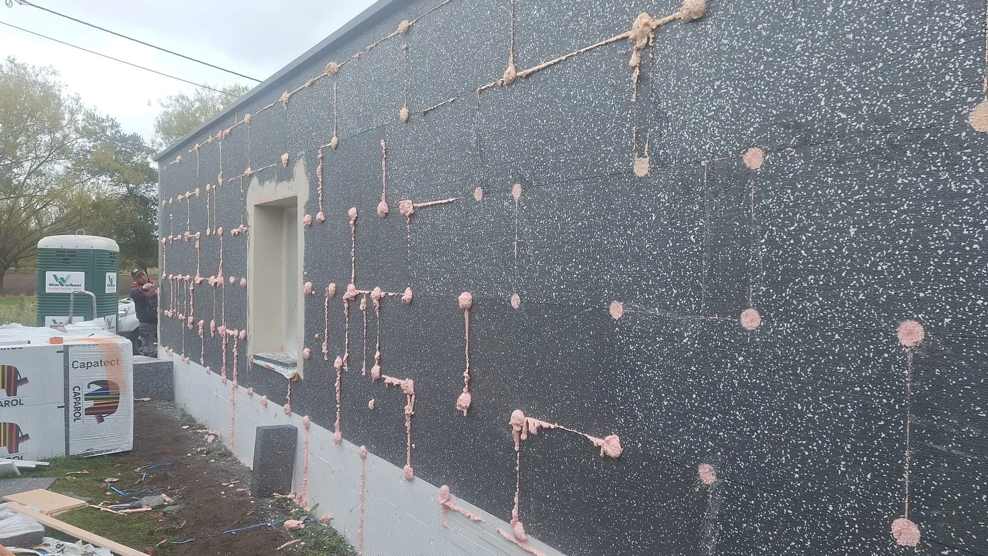 Construction site showing the exterior wall of a building with black foam insulation installed and pink spray foam insulation applied at the joints. There are construction materials and workers in the background.
