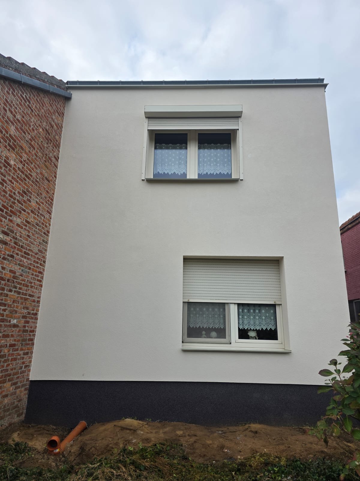 A two-story white residential building with two windows, one on each floor. The upper window has lace curtains and a roller shutter, while the lower window has lace curtains and a small decorative item on the sill. The building is next to a brick wal