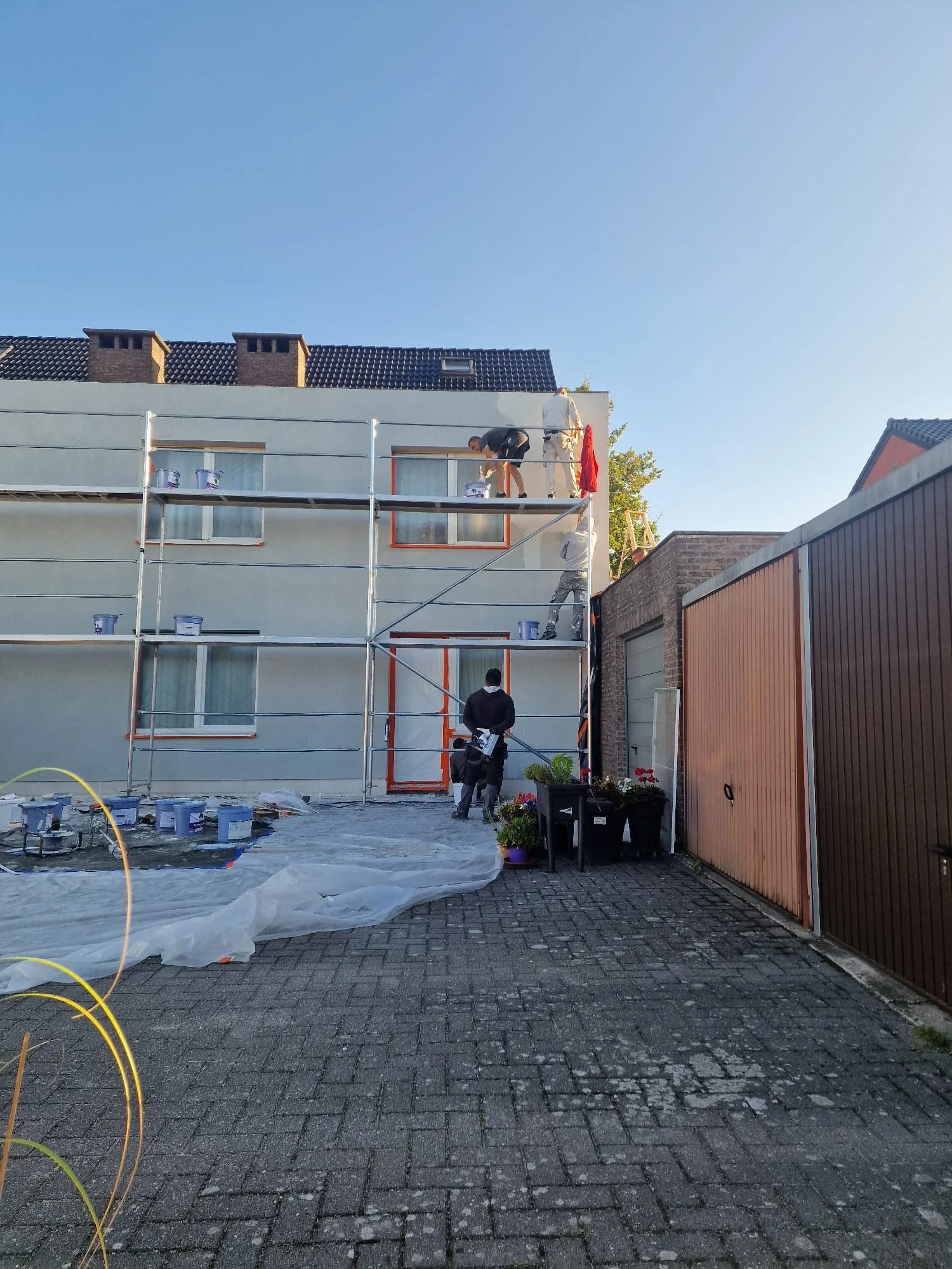 Workers painting the exterior of a multi-story residential building, with scaffolding set up in front and various painting supplies on the ground.
