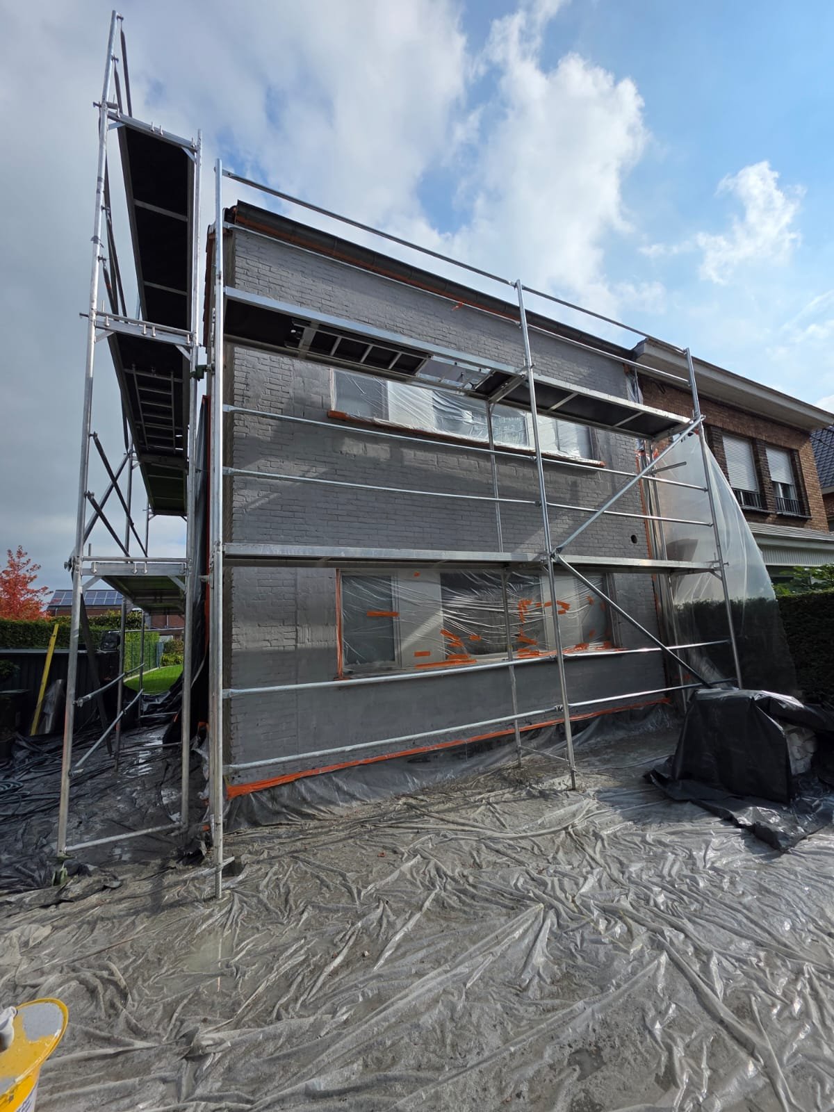 House under renovation with scaffolding, plastic covering, and tarps on the ground, with a cloudy sky overhead.