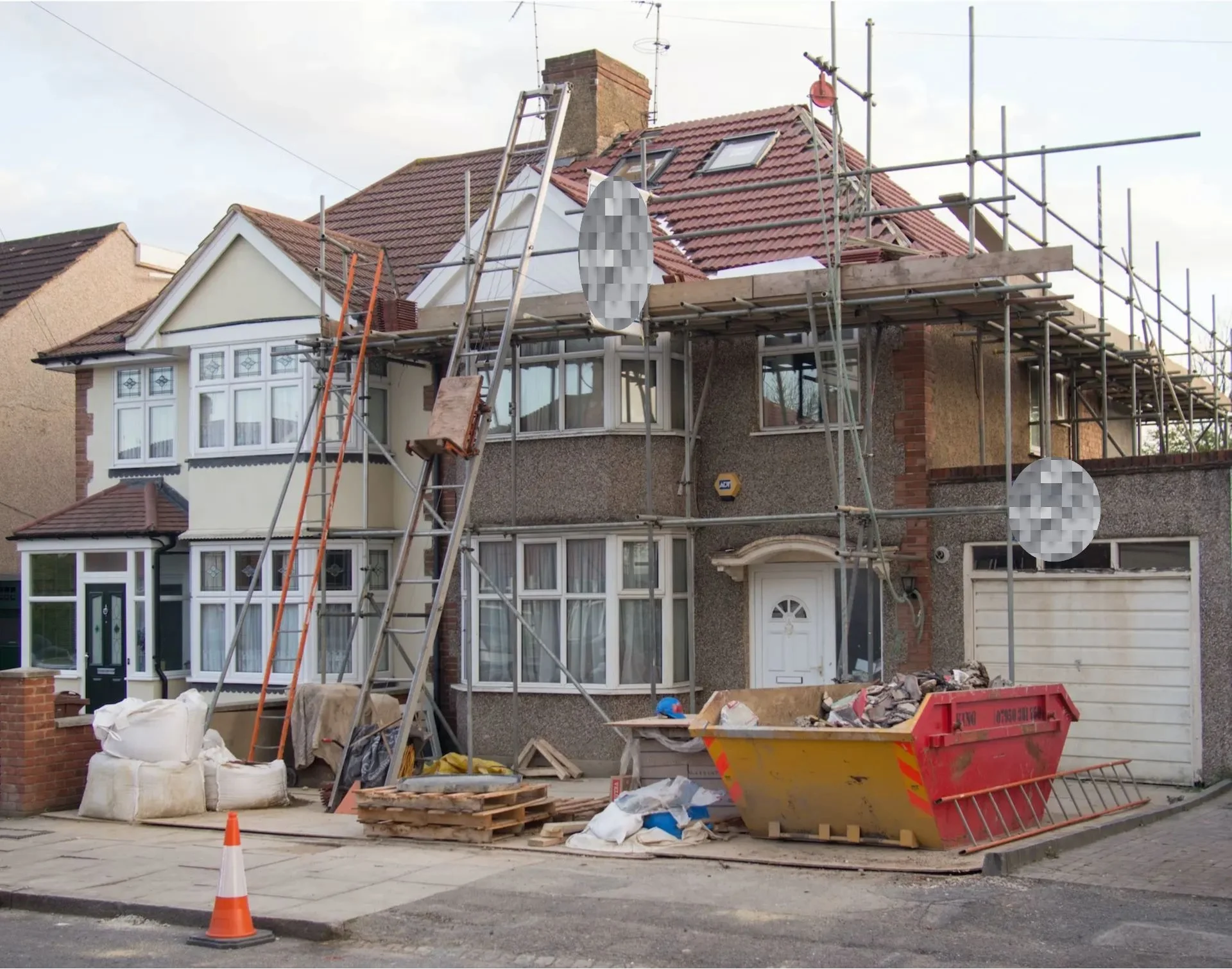 A two-story house under renovation with scaffolding around the roof and front, a construction dumpster filled with debris in the driveway, building materials and tools on the ground, and orange safety cones on the street.