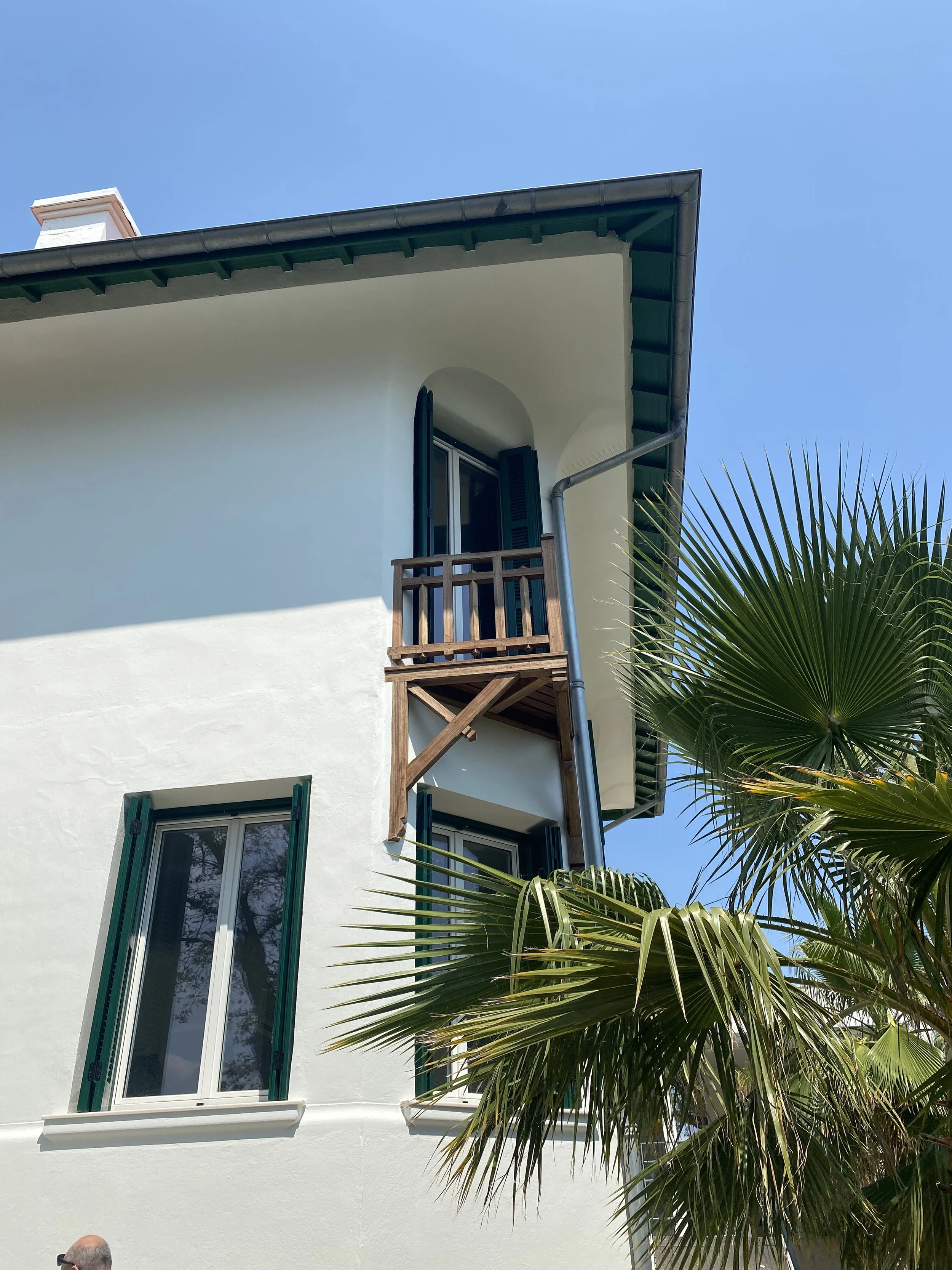 White multi-story house with dark green shutters, small balcony with wooden railing, and a palm tree in front, set against a clear blue sky.