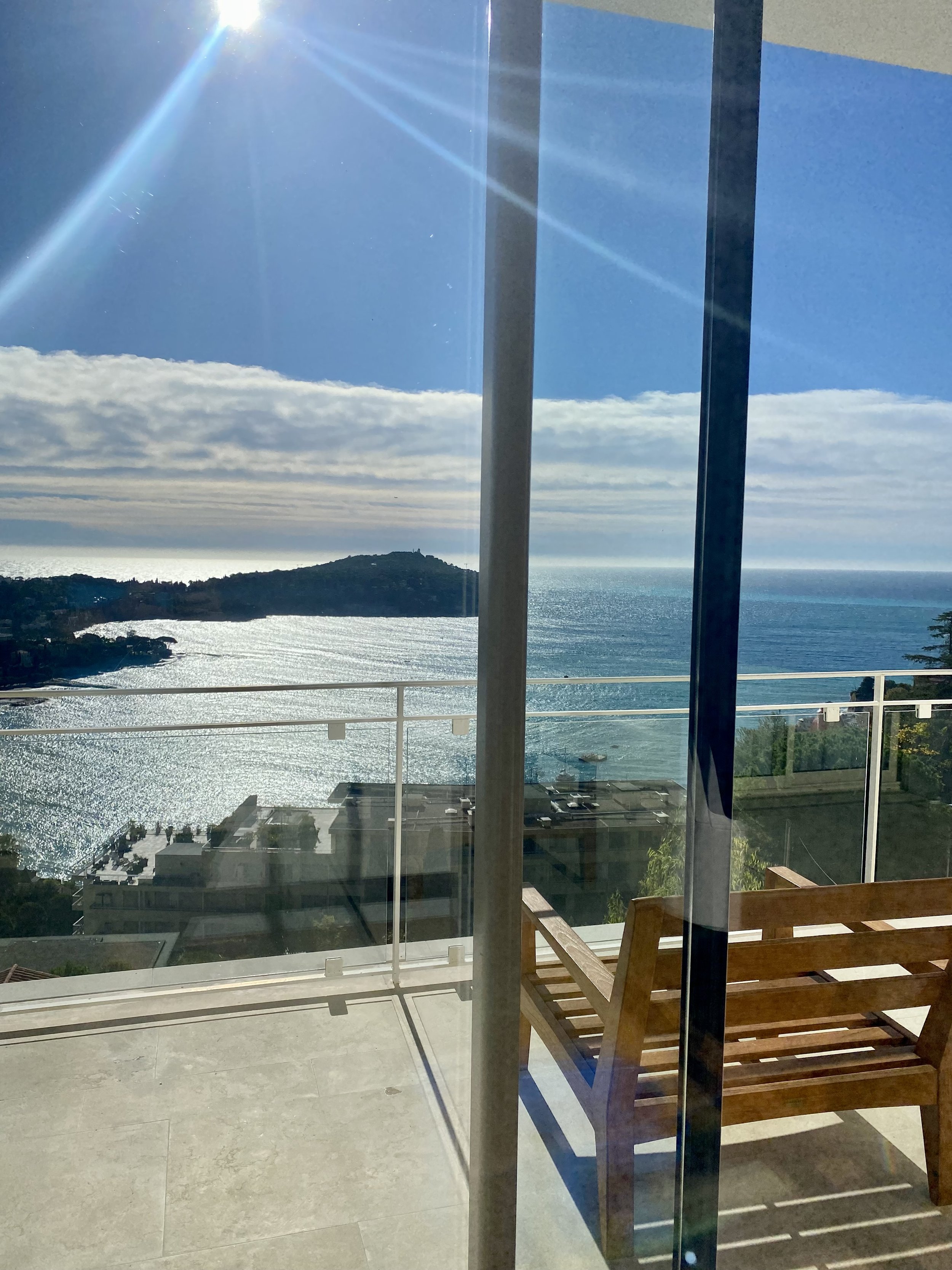 A balcony with a wooden bench overlooking a large body of water, with hills and partly cloudy sky in the distance, seen through a glass door with sunlight glare.