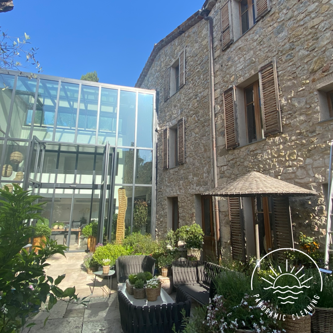 A cozy outdoor patio area with black outdoor furniture, potted plants, and a stone building with wooden shutters. There's also a part of a modern glass extension with a door open to the patio, under a blue sky.