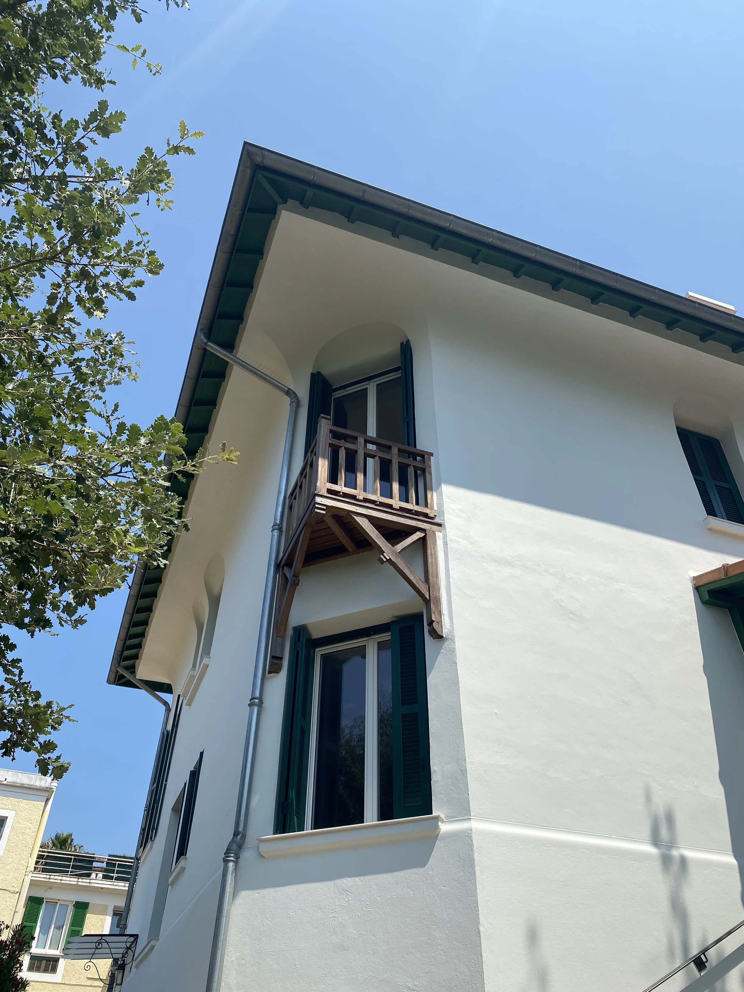 A white building with blue shutters and a small wooden balcony on the second floor, with a clear blue sky above and some green tree branches on the left.