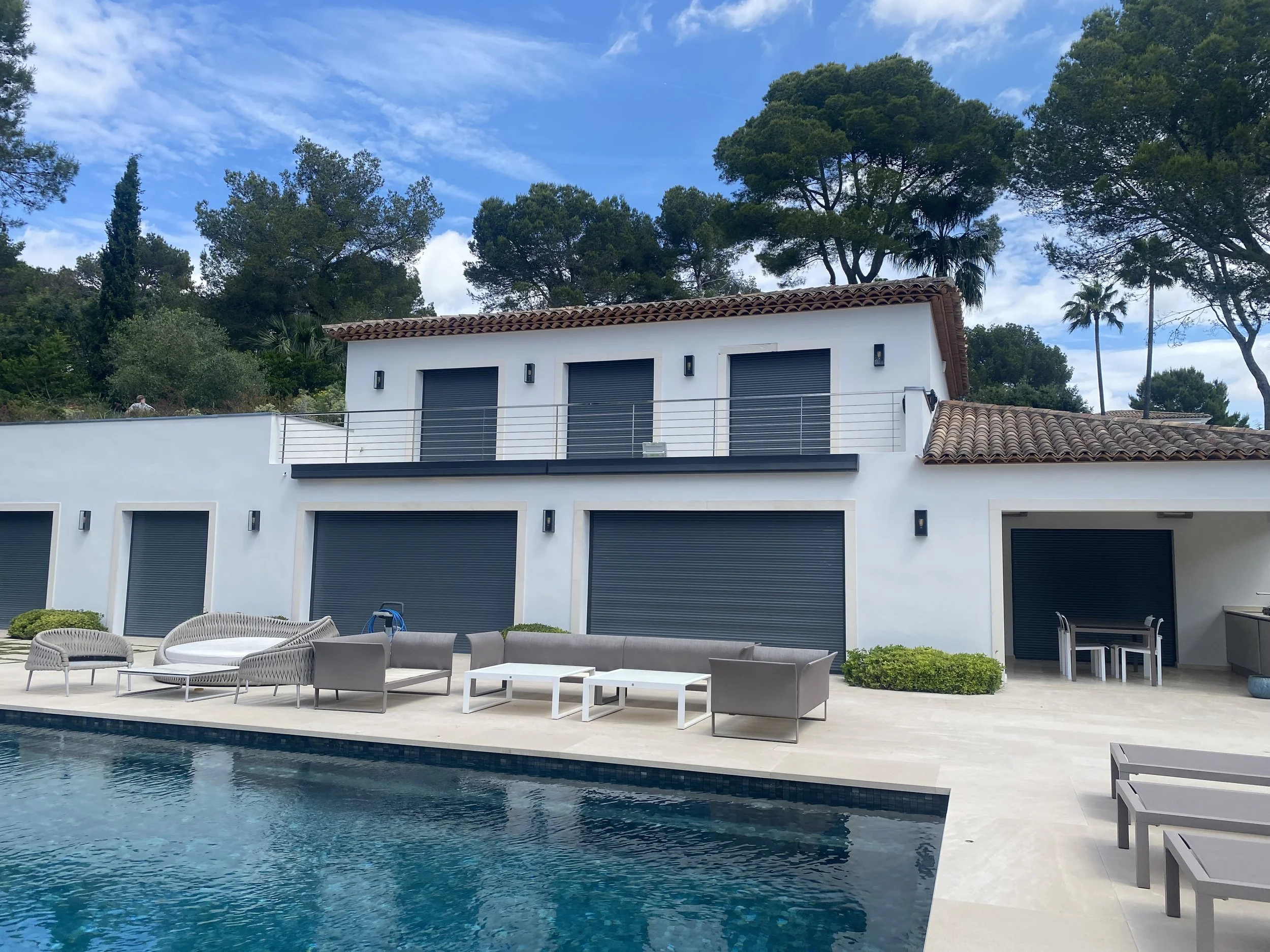 Modern white house with closed black shutters, outdoor seating by a swimming pool, and surrounded by green trees under a partly cloudy sky.