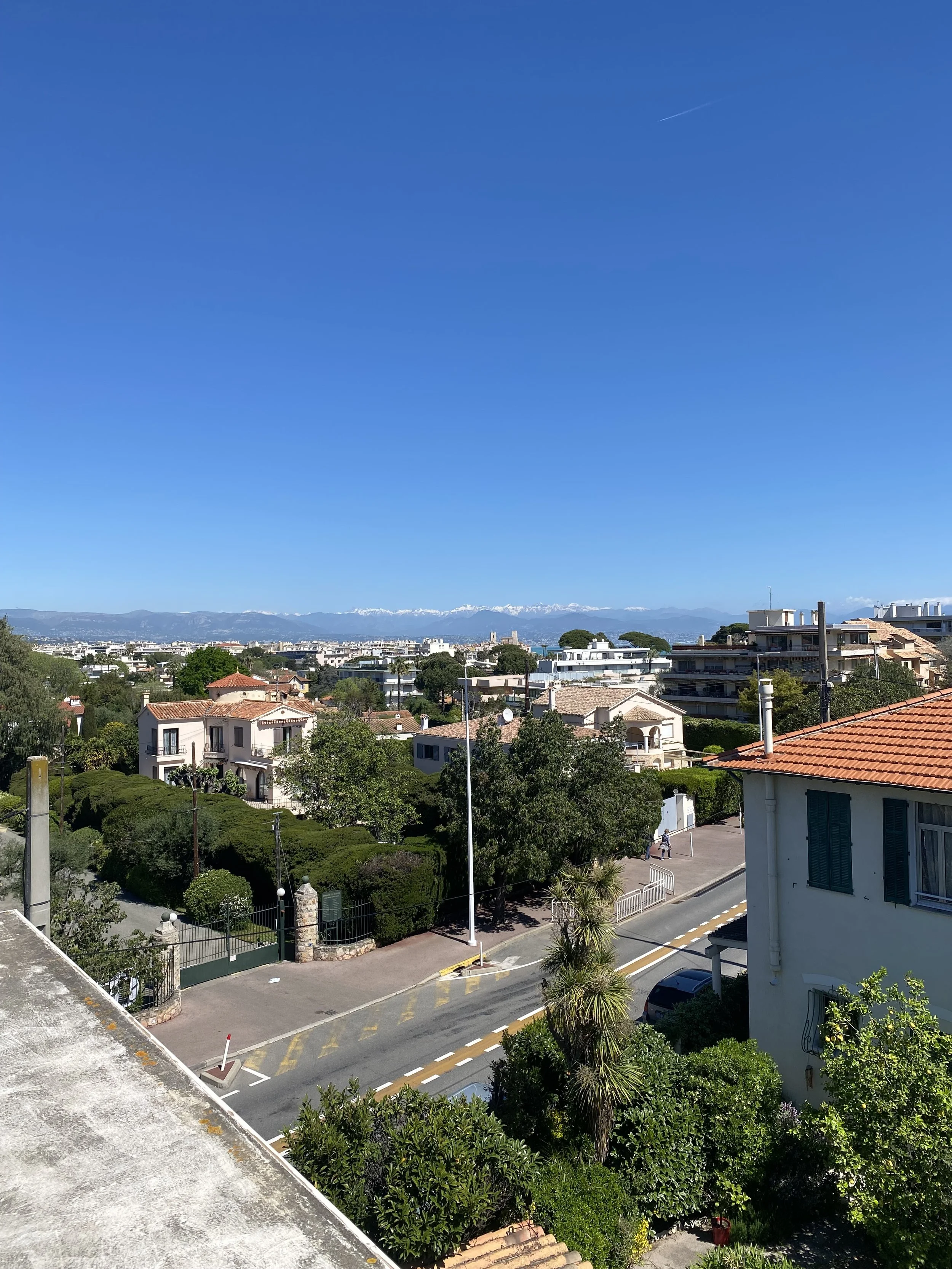 Scenic view of a residential neighborhood with trees, houses with red-tiled roofs, and a distant mountain range with snow-capped peaks under a clear blue sky.