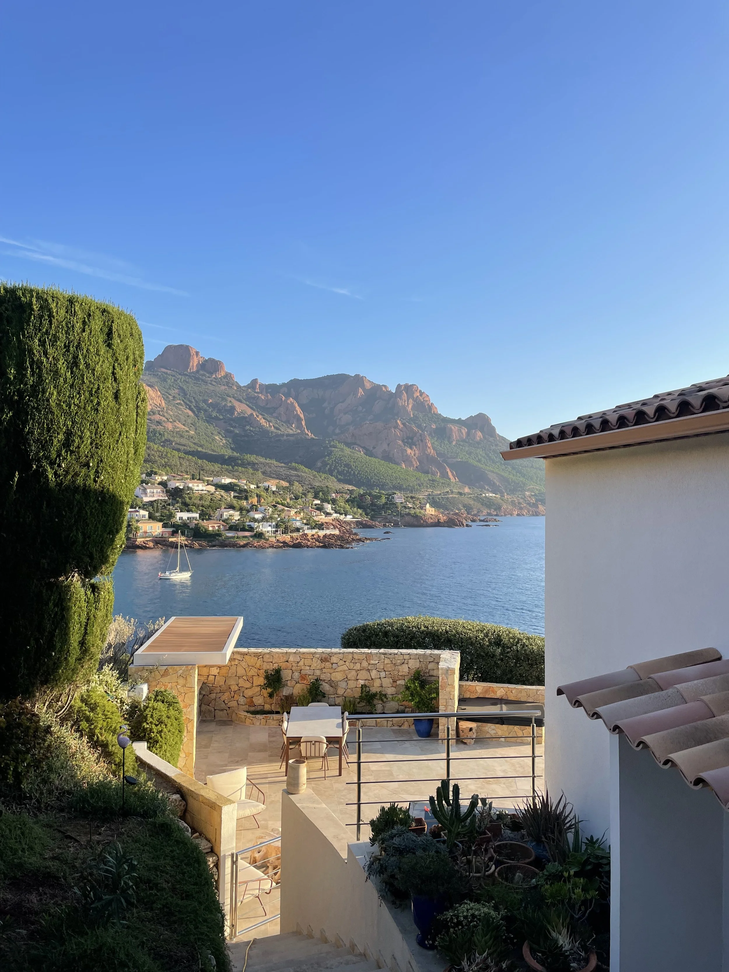 Coastal view with mountains, water, a sailboat, and houses on the hillside, seen from a patio with plants and outdoor furniture.