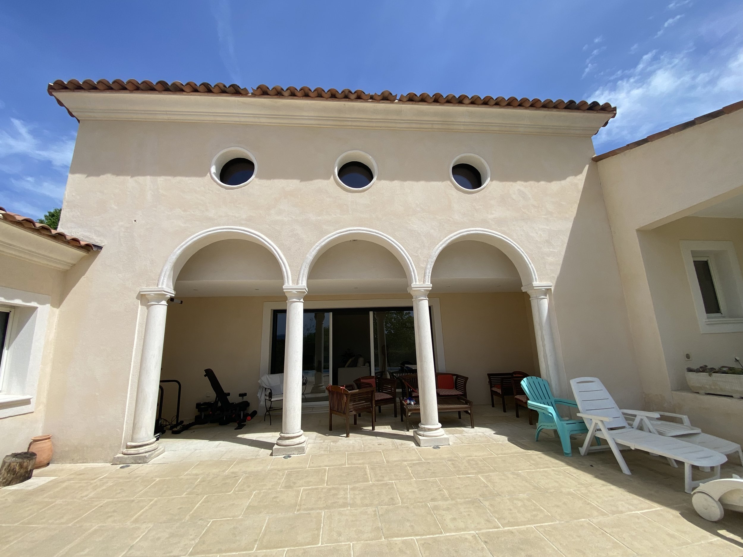 Outdoor patio with arched columns, lounge chairs, and patio furniture in front of a beige stucco house with circular windows, under a blue sky.