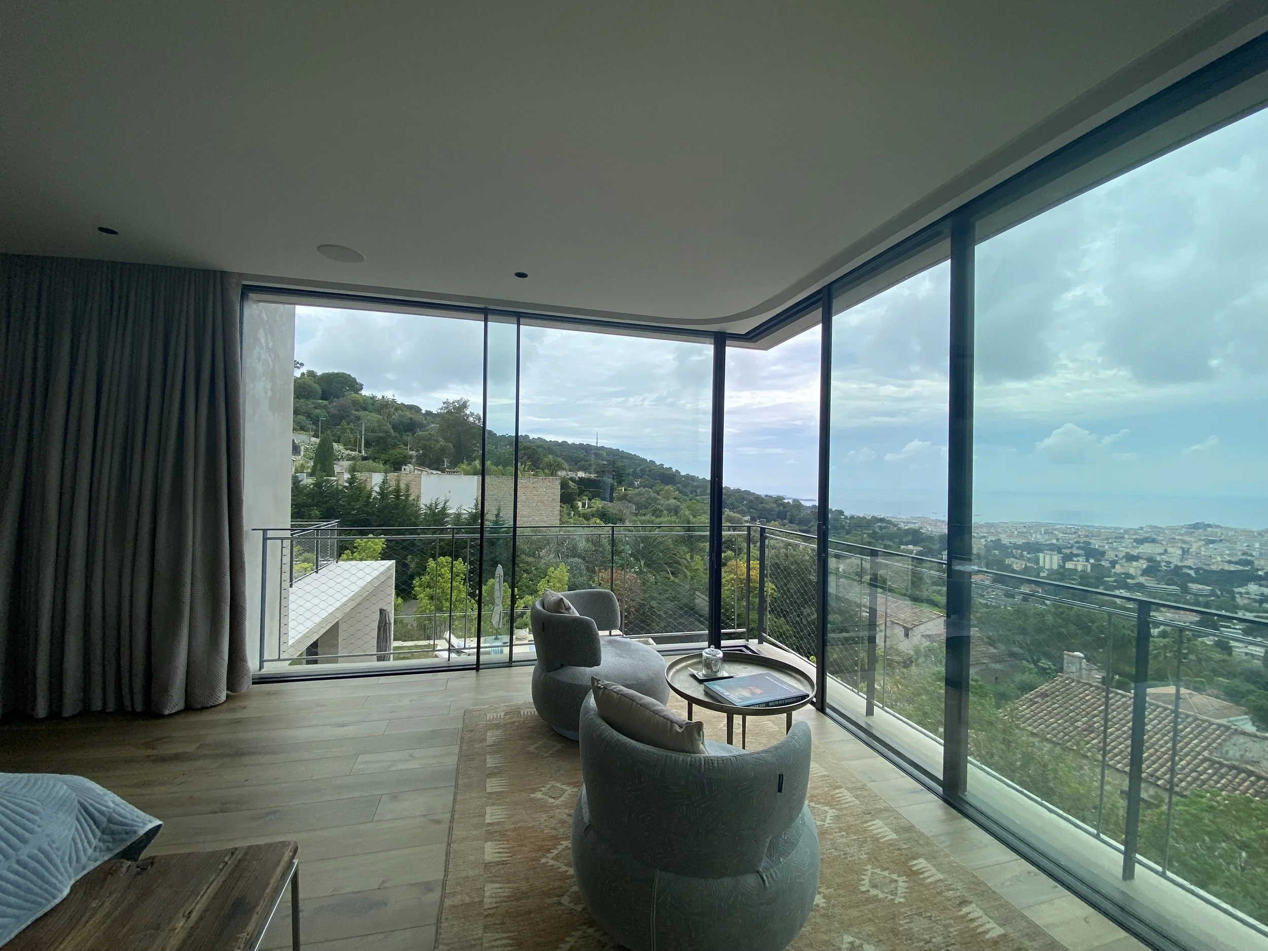 Living room with large floor-to-ceiling windows showing a hilly landscape and cityscape view, two modern armchairs, a small round coffee table with magazines, a rug, and beige curtains on the left.