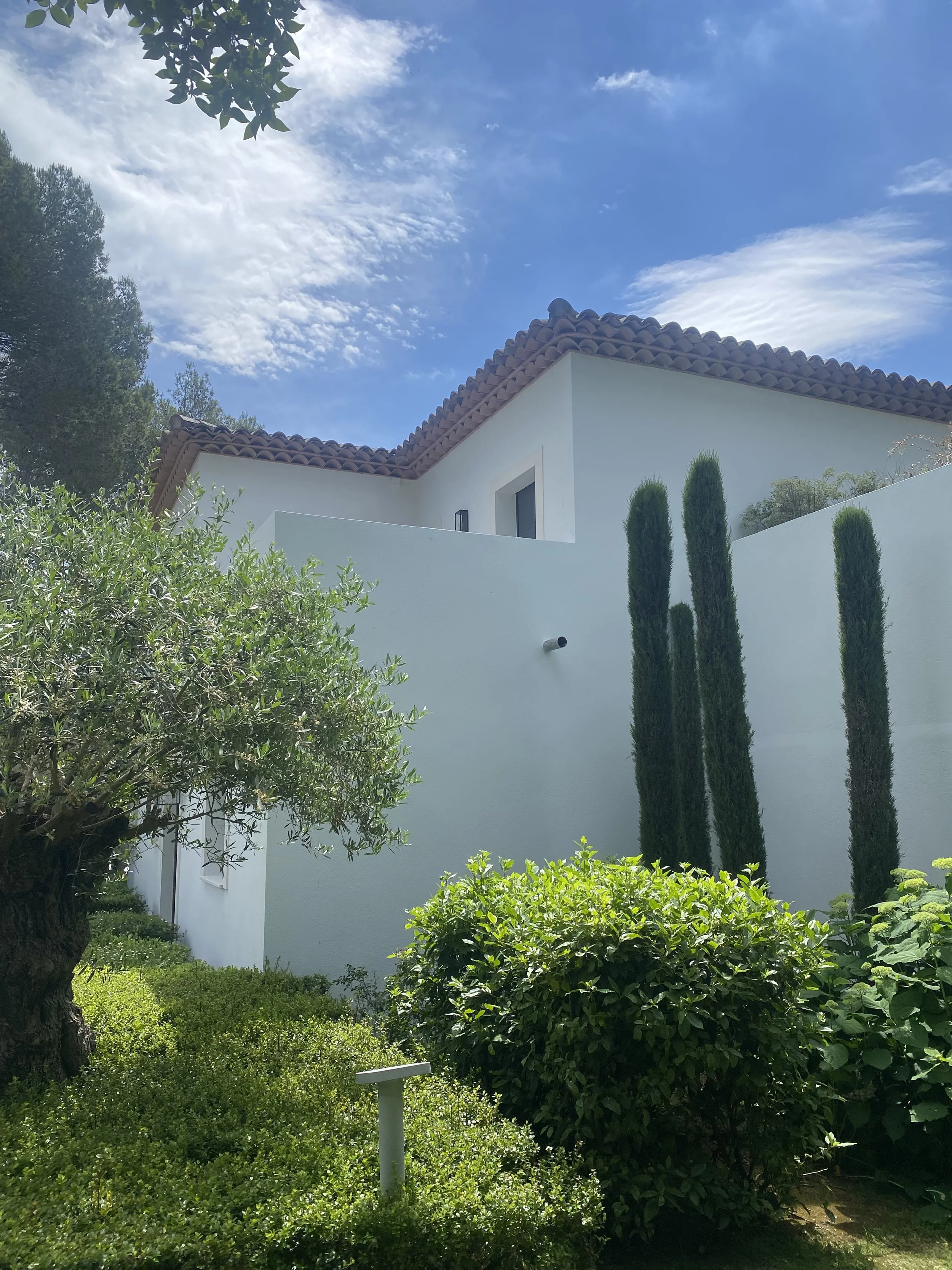 A modern white house with a red tile roof, surrounded by green bushes and tall cypress trees, under a blue sky with scattered clouds.
