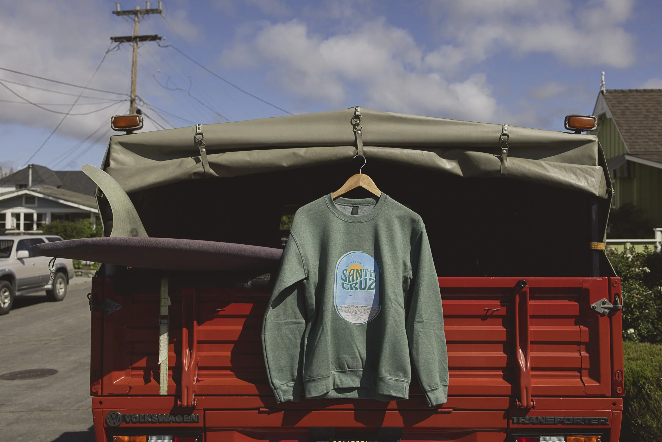 Green sweatshirt with a Santa Cruz logo hanging on a wooden hanger from the top of a red Volkswagen Transporter truck's tailgate, with two surfboards on the truck's back, parked on a suburban street under a partly cloudy sky.
