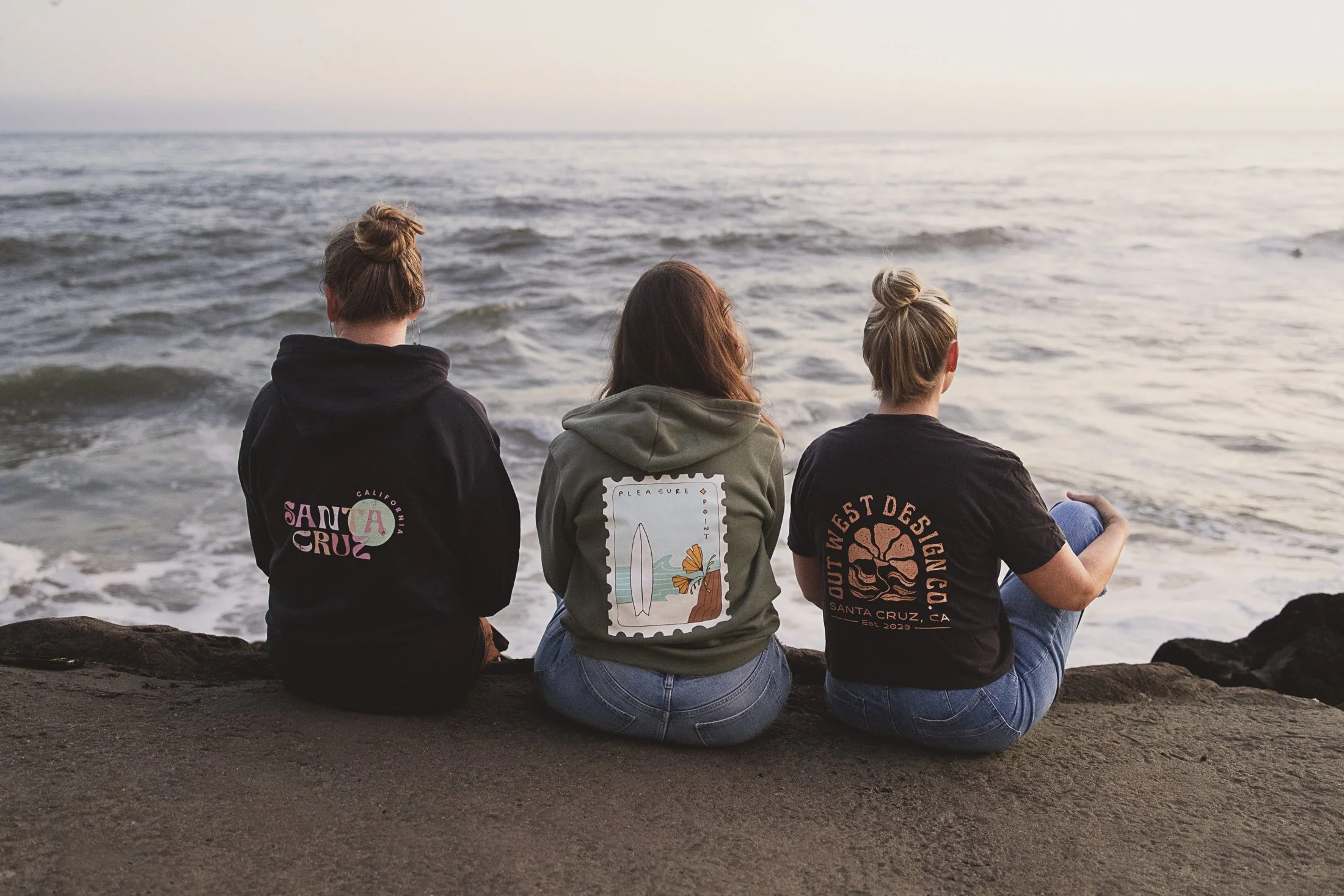 Three women sit on the rocks facing the ocean and watch the sunset at the beach.