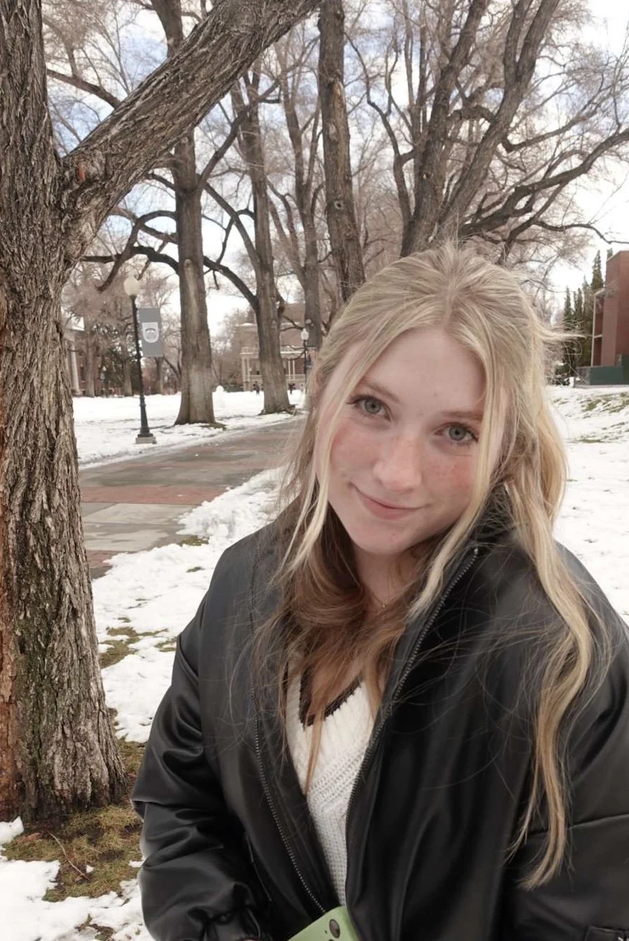 A young woman with blonde hair and blue eyes, wearing a black jacket and a white sweater, smiling outdoors in a snowy park with trees and brick buildings in the background.