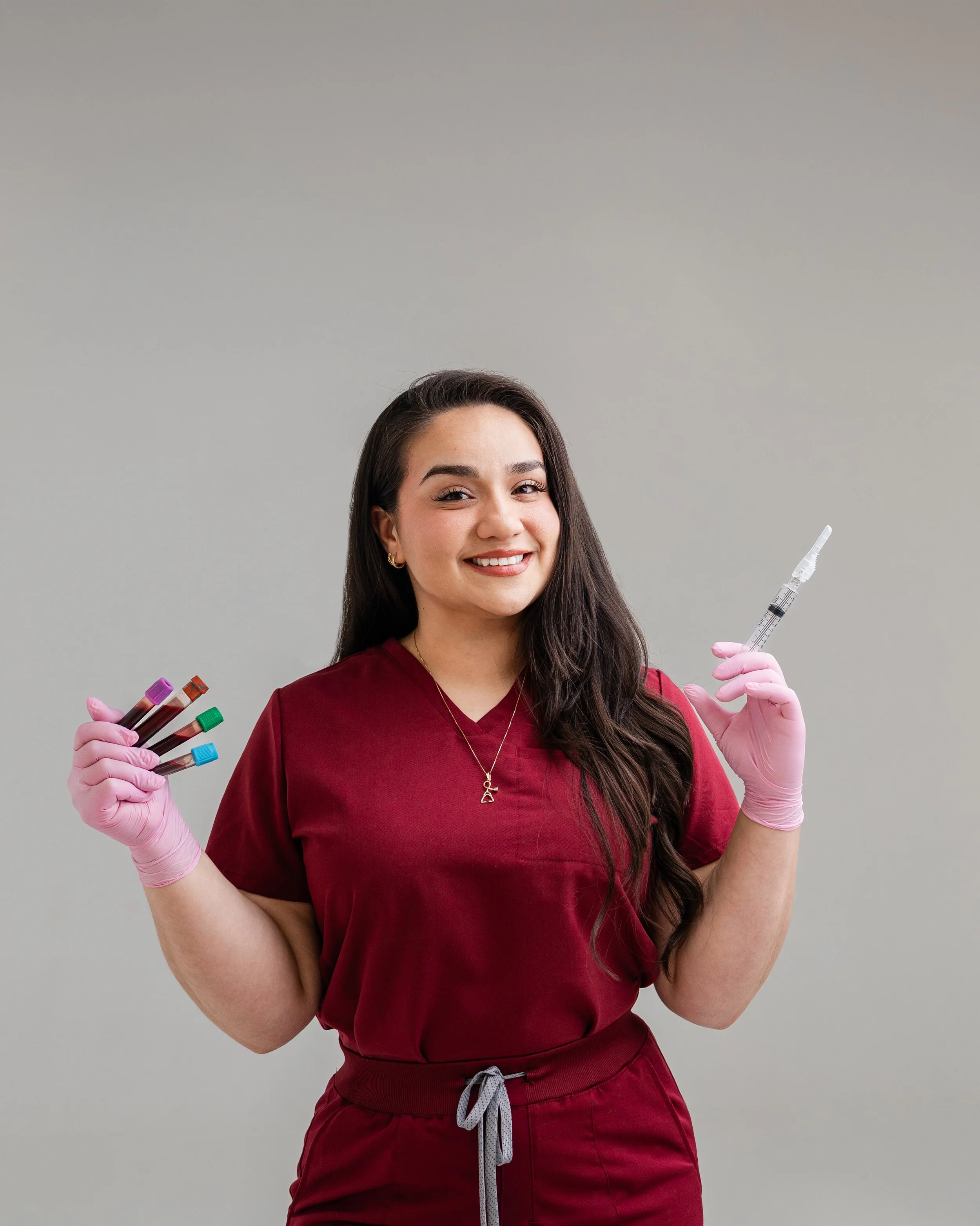 A smiling young woman with long dark hair wearing burgundy medical scrubs and pink gloves is holding several blood sample vials in her left hand and a syringe in her right hand, standing against a plain light gray background.