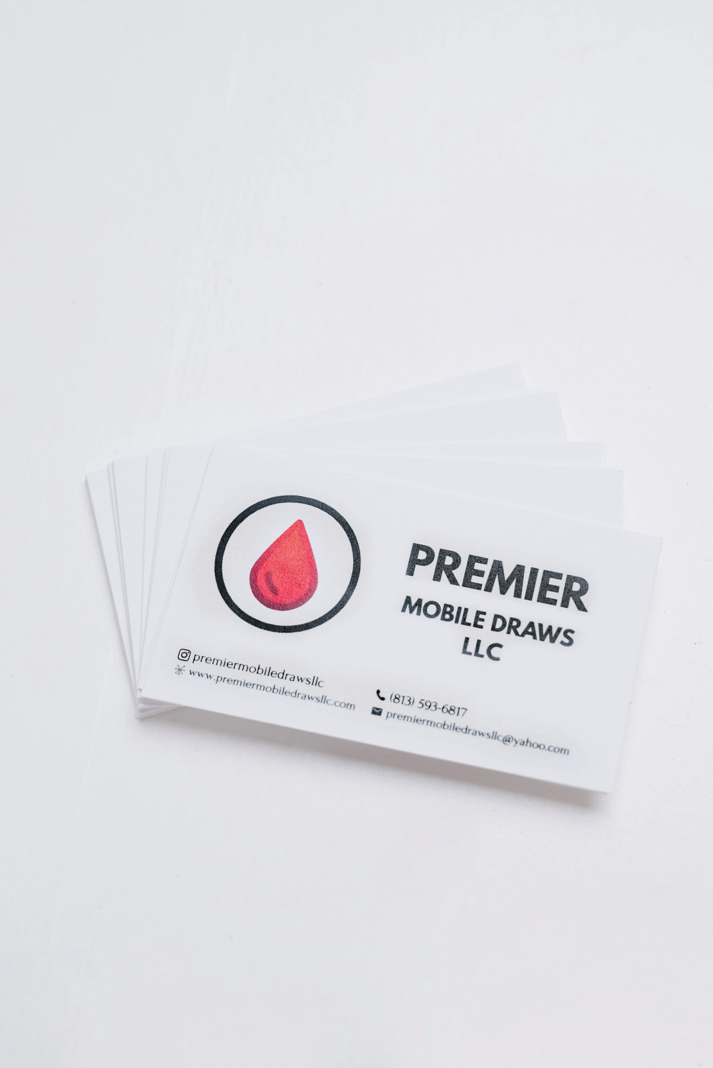 Business cards for Premier Mobile Draws LLC stacked on a white surface, featuring a logo with a red drop inside a black circle, and contact information.