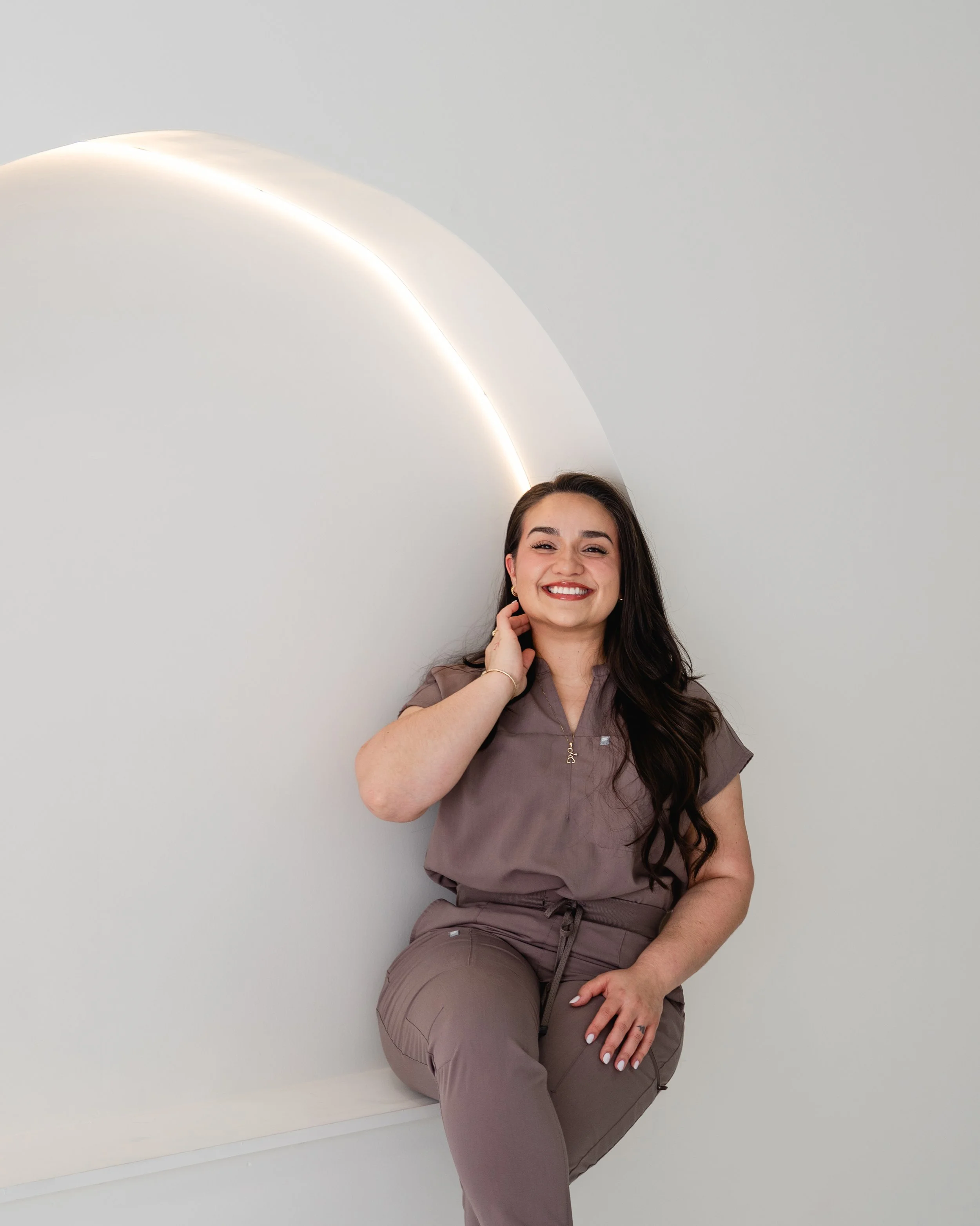 A smiling woman with long dark hair, wearing medical scrubs, sitting against a white wall with curved lighting, touching her neck with her right hand.